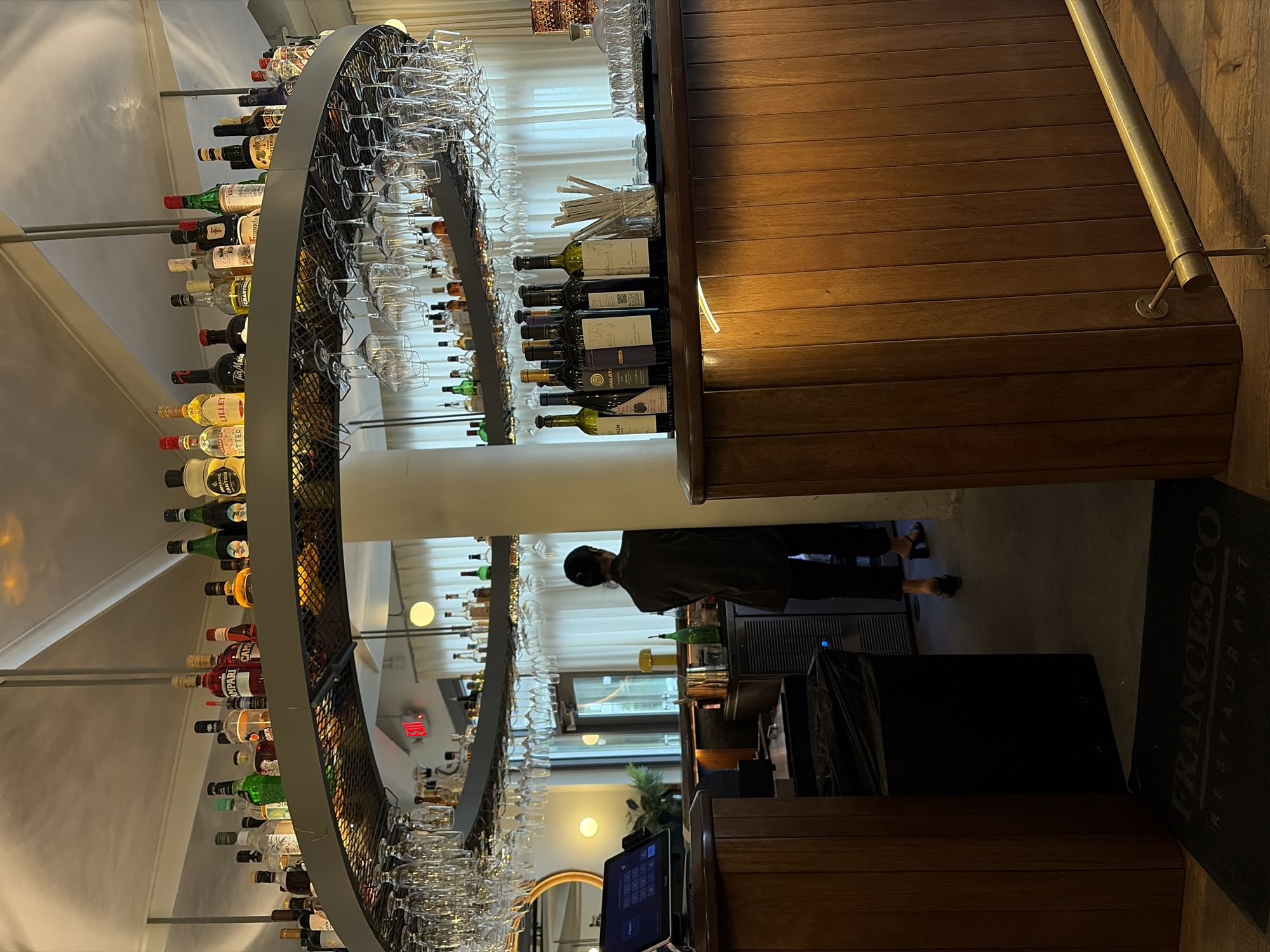 This is an interior shot of Francesco Restaurant in Coral Gables, Florida. A semicircular bar, laden with liquor bottles and glassware, dominates the left side of the frame. The bar features a metal frame with shelves to display the alcoholic beverages and glassware. The space behind the bar is partially obscured by a pillar. The restaurant's interior design is defined by dark wood paneling. A man is seen standing behind the bar. A black mat labeled "FRANCESCO RESTAURANT" lies on the floor.