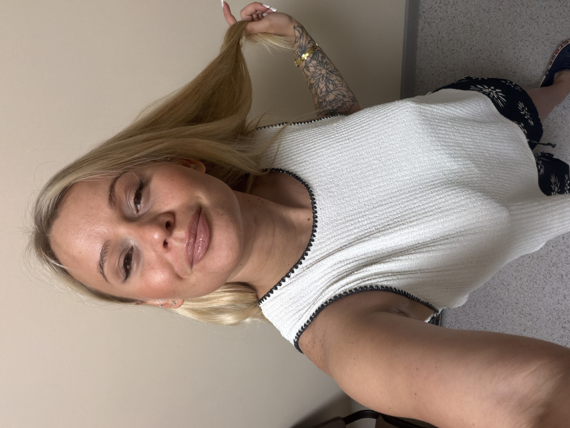 A woman with blonde hair is posing for a selfie, holding her hair in her right hand. She is wearing a white sleeveless top with black trim and dark patterned shorts. She has tattoos on her left arm and is wearing a gold bracelet. The background appears to be an indoor setting with a beige wall and a grey tiled floor. There is no indication of the location being in Sarapul, Russia.