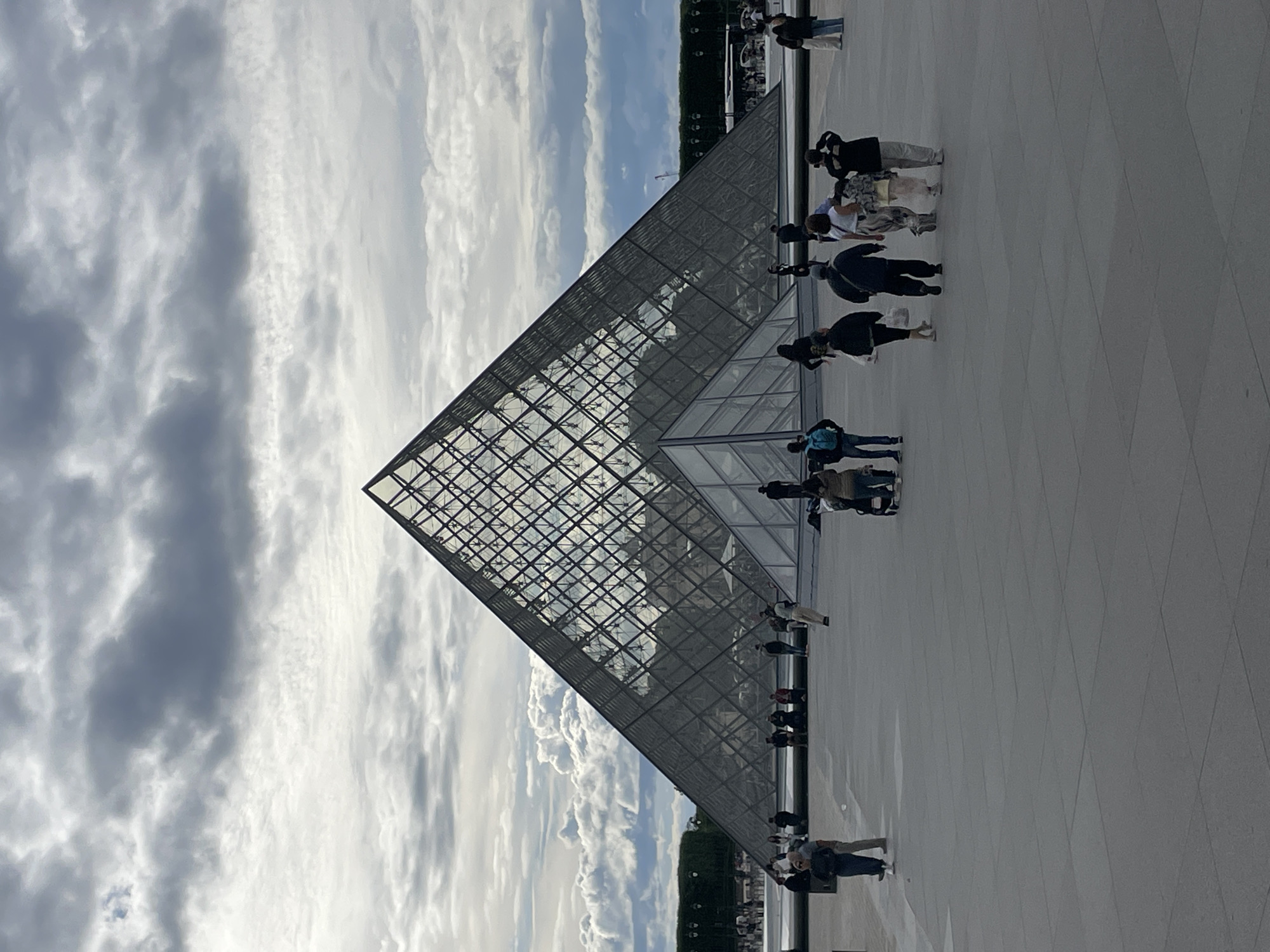 The image depicts the Louvre Pyramid in Paris, France. It is a cloudy day, with dramatic grey and white clouds filling the sky. The glass pyramid is the central focus, its geometric structure reflecting the sky and surrounding architecture. Several people are visible in the foreground and midground, walking and standing on the wide, paved plaza. Some are looking towards the pyramid, while others are engaged in conversation or moving about. The plaza itself is a large, open space with a grey, stone-like surface. In the background, there are trees and buildings, suggesting the expansive grounds of the Louvre Museum. The overall impression is one of a busy public space on a somewhat overcast day.