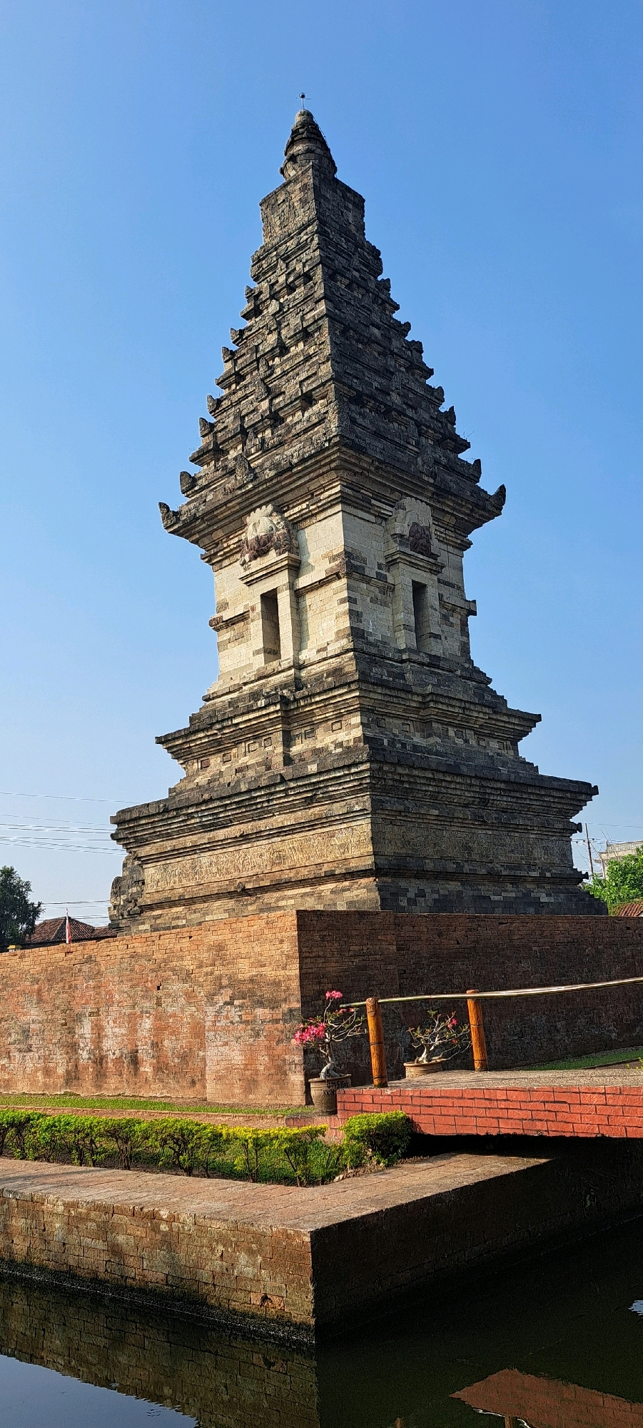 The image depicts the Candi Wates temple complex in Kediri, East Java, Indonesia, under a clear blue sky. The central subject is a multi-tiered, stone-built pagoda-style temple, constructed from weathered grey and light brown stone blocks. The temple features intricate carvings, small rectangular window-like openings, and decorative roof elements. It stands on a raised platform, surrounded by a red brick wall. In the foreground, a body of dark water reflects the structures and the sky. A narrow brick pathway and a small bridge with wooden railings are visible, leading towards the temple. To the left of the temple, a section of the brick wall has a few potted plants, including one with bright pink flowers. The overall environment appears to be a well-maintained historical site, with some greenery and shadows cast by the structures. There are no people visible in the image.