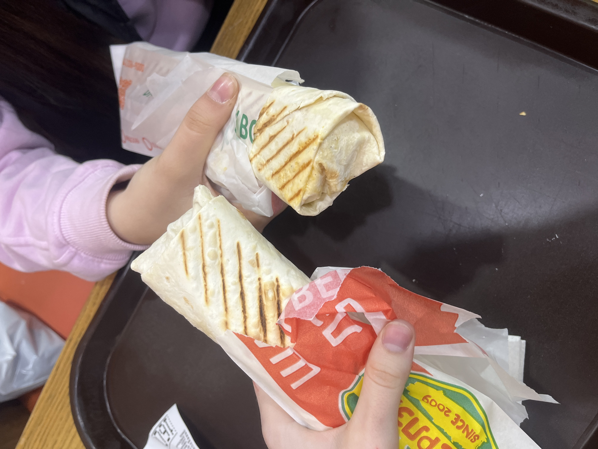 Two hands are holding shawarma wraps in paper wrappers. One wrapper is red and white and the other is white with green lettering. The background is a dark tray on top of a wooden table. A person in a pink sweatshirt is partially visible. Based on the green and yellow wrappers that say "Piters", this image was likely taken in Saint Petersburg, Russia.