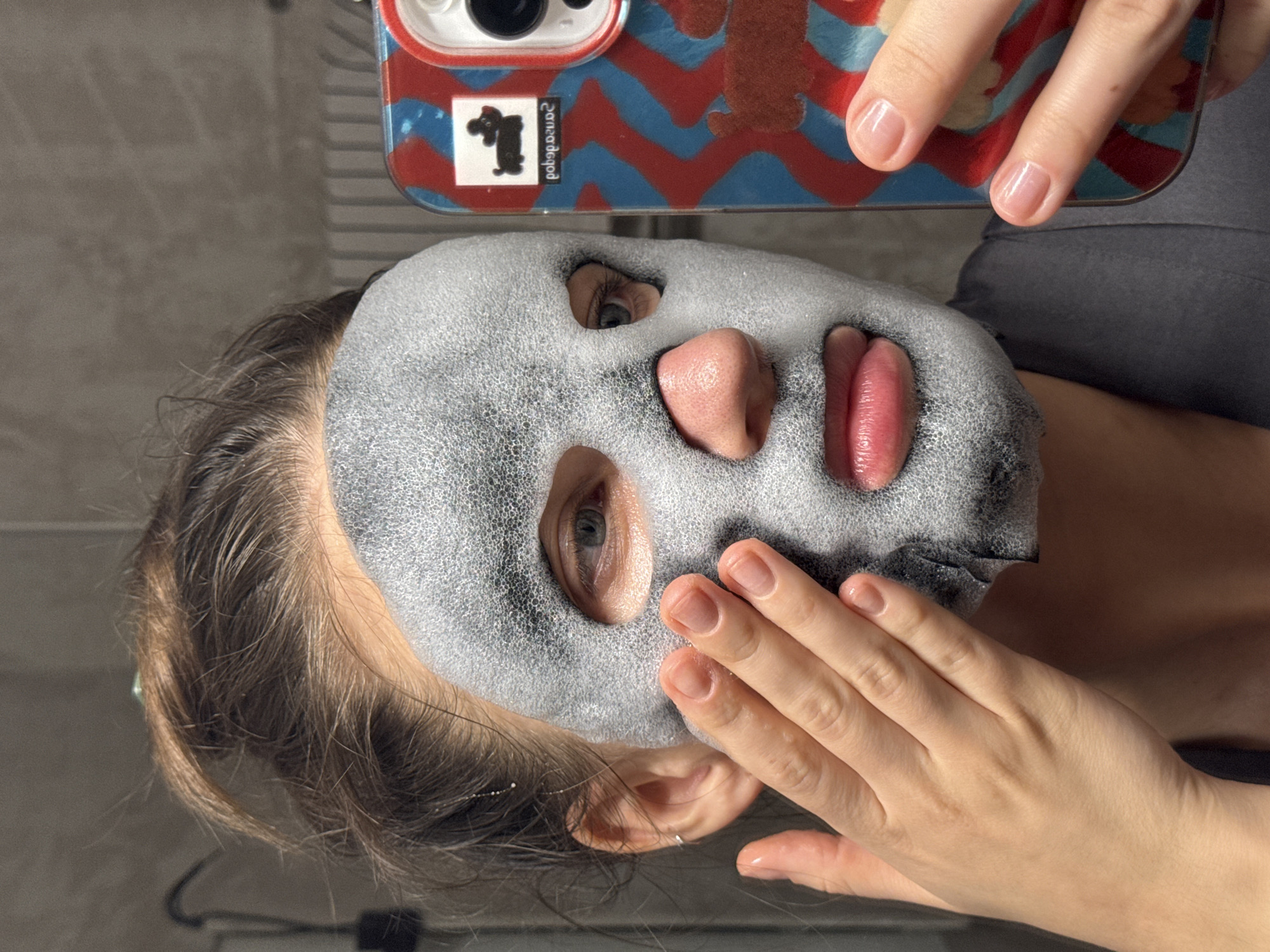 A mirror selfie captures a person, likely a woman, engaged in a skincare routine while wearing a grey, foamy bubble face mask. The mask covers the majority of her face, with openings for her eyes, nose, and mouth, revealing her open eyes and slightly parted lips. Her brown hair is visible framing her face.
The person's right hand (upper part of the image) is holding a smartphone, indicating the selfie is being taken into a mirror. The phone case is distinctly patterned with red and blue waves or zigzags and features a white sticker with a black cartoon dog figure. Text on this sticker is visible and mirrored, appearing as "2020dogod" or "ZonoDogod" (likely "dogod 2020" when not mirrored). Her left hand (lower right side of the image) is gently resting on her cheek, near the mask. Her fingernails are natural and neatly manicured.
The background suggests an indoor setting, possibly a bathroom, with a grey, textured or tiled wall visible behind her. The lighting is soft and even, typical of an indoor environment. The person's expression is neutral, primarily conveyed through her eyes due to the mask. The overall scene depicts a personal moment of self-care.