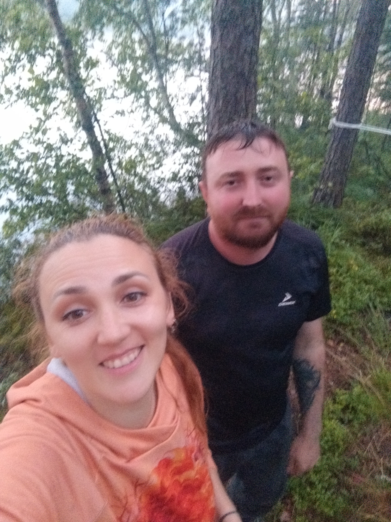 Two individuals, a woman and a man, are pictured in a selfie-style photograph. The woman is in the foreground, on the left, wearing an orange hooded sweatshirt. She has reddish-brown, curly hair and is smiling. The man is behind and to the right of the woman, wearing a black t-shirt. He has short brown hair, a beard, and a tattoo on his left arm. They appear to be in a natural setting, possibly a forest or park, with trees and greenery visible in the background. A white rope is strung between two trees behind the man. The lighting suggests it may be late afternoon or early evening. The image is a close-up of the two people, with the environment serving as a backdrop. The location is identified as Tarasovskoe, Russia.