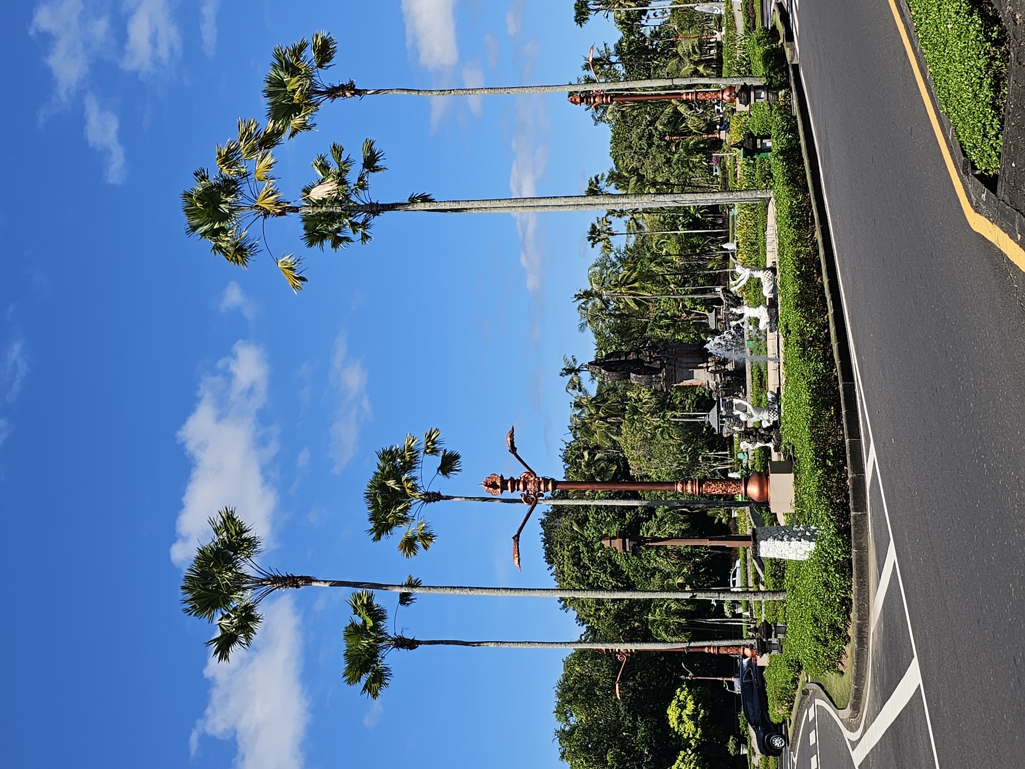 The image depicts a street scene in Kuta Selatan, Indonesia. A paved road with a yellow line marking the edge is visible on the right side. Adjacent to the road is a landscaped area featuring manicured green hedges, tropical trees, and Balinese decorative elements, including statues and lampposts. Several tall palm trees are prominently displayed against a bright blue sky with scattered white clouds. In the background, dense tropical foliage and a glimpse of a Balinese structure or monument can be observed. A car can be seen parked along the edge of the road. The overall impression is that of a well-maintained and tropical urban landscape.