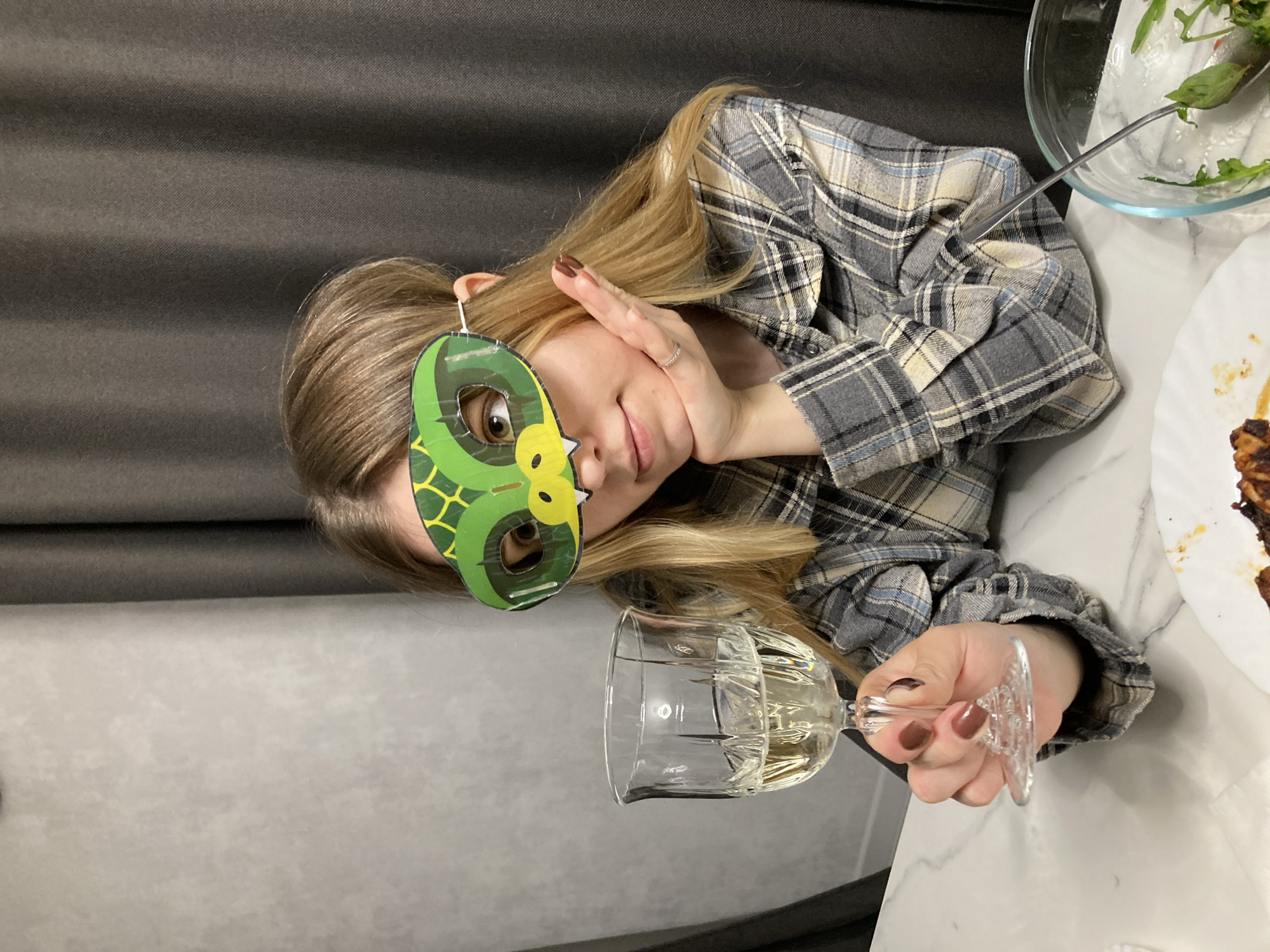 A blonde woman with fair skin, wearing a gray plaid shirt and a green and yellow mask shaped like a reptile's head, holds a clear wine glass filled with a light-colored liquid. Her left hand is near her face, with a ring visible on one of her fingers. The background includes a gray fabric wall, a white marble-patterned table, a glass bowl with salad, and a white plate with food. The location is Chaikovsky, Russia.
