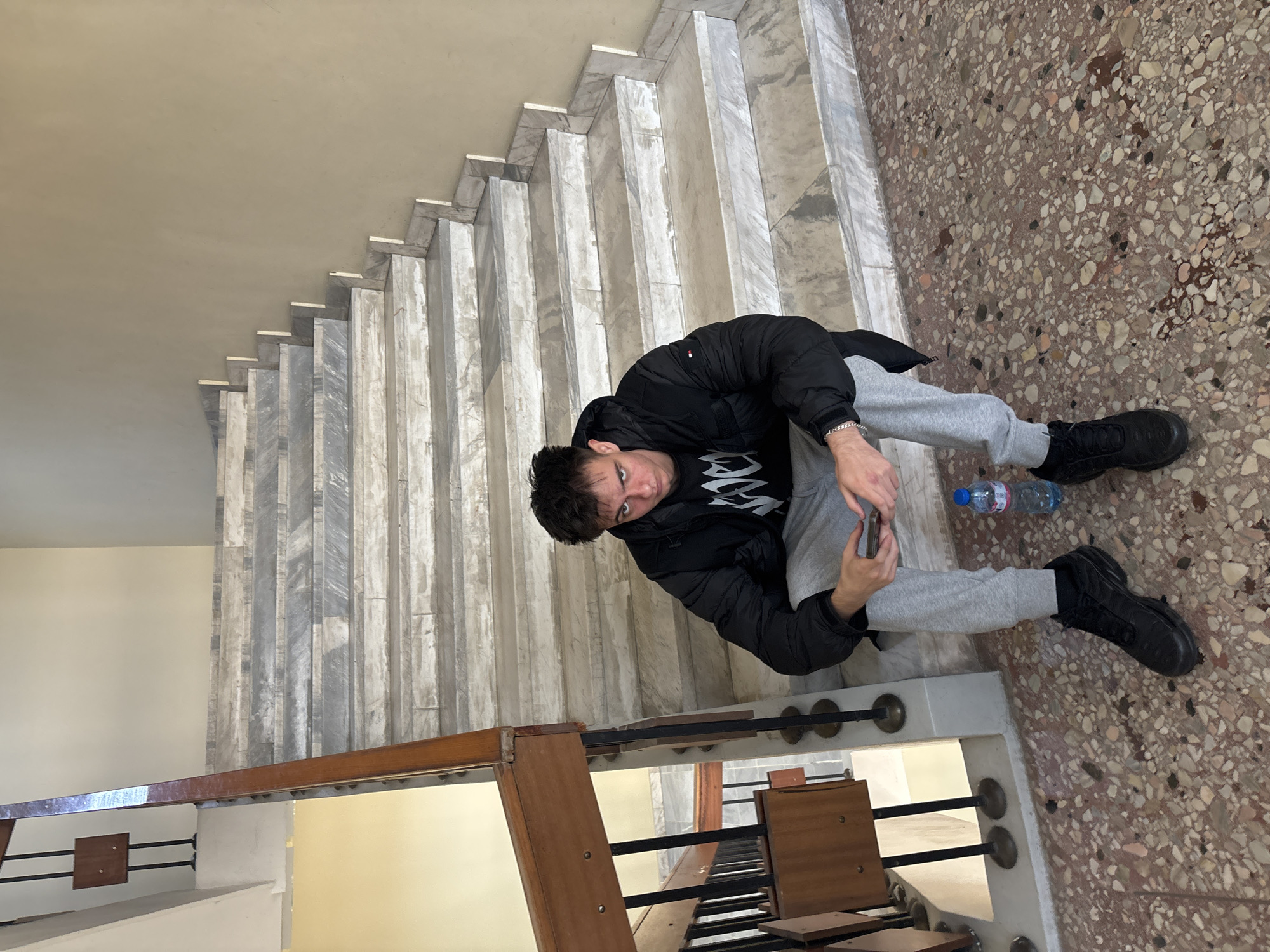A young man in a black jacket and grey sweatpants is sitting on the steps of a marble staircase in Plovdiv, Bulgaria, possibly within an architectural building or structure. He is holding a phone and looking directly at the camera. Next to him is a plastic water bottle. The floor is a terrazzo style with a mosaic of various colors. The staircase is adjacent to a wooden railing, and beneath the railing are some benches. The overall environment is indoor, presumably within a public building.