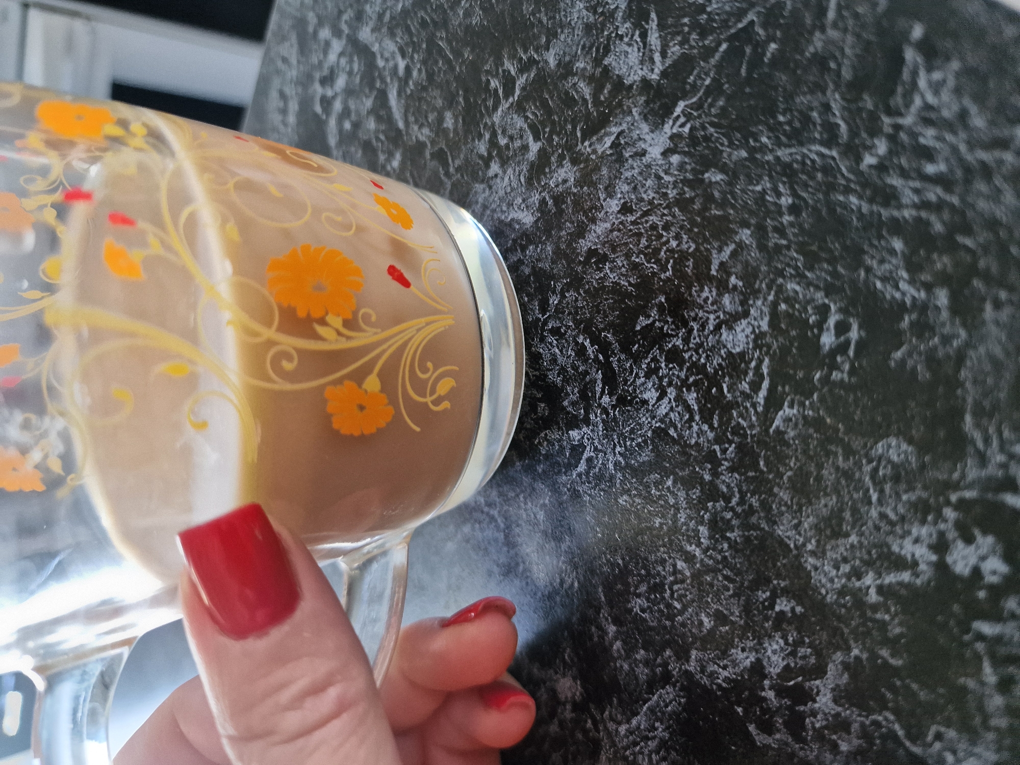 A close-up shot shows a hand with red nail polish holding a transparent mug filled with a light brown beverage. The mug has a decorative pattern of orange flowers and yellow swirls. The background is a dark, textured surface, possibly a countertop. The scene is devoid of any specific location markers within Kursavka, Russia.
