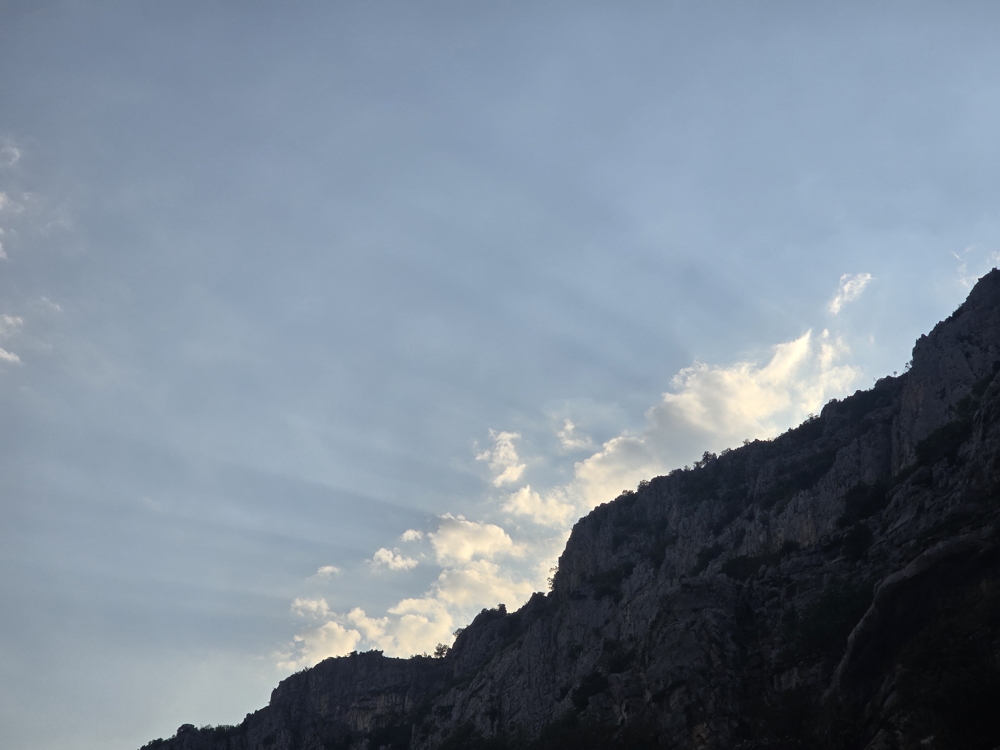 The image shows a dramatic, silhouetted mountain range against a pale blue sky with scattered clouds. The sun is not visible, but light rays fan out from behind the clouds, illuminating some of the cloud edges and casting a soft light. The rocky slopes of the mountains are steep and rugged, with sparse vegetation visible along the crest. The overall impression is one of rugged natural beauty.
This scene is likely the Skadar Lake National Park, in Montenegro, a region known for its dramatic mountain landscapes and proximity to Podgorica. While specific landmarks within the park are not identifiable, the terrain is consistent with the karst topography found in the area. There are no people, animals, or man-made structures visible in the image. The environment is natural and untamed, with the focus on the geological formations and atmospheric conditions.