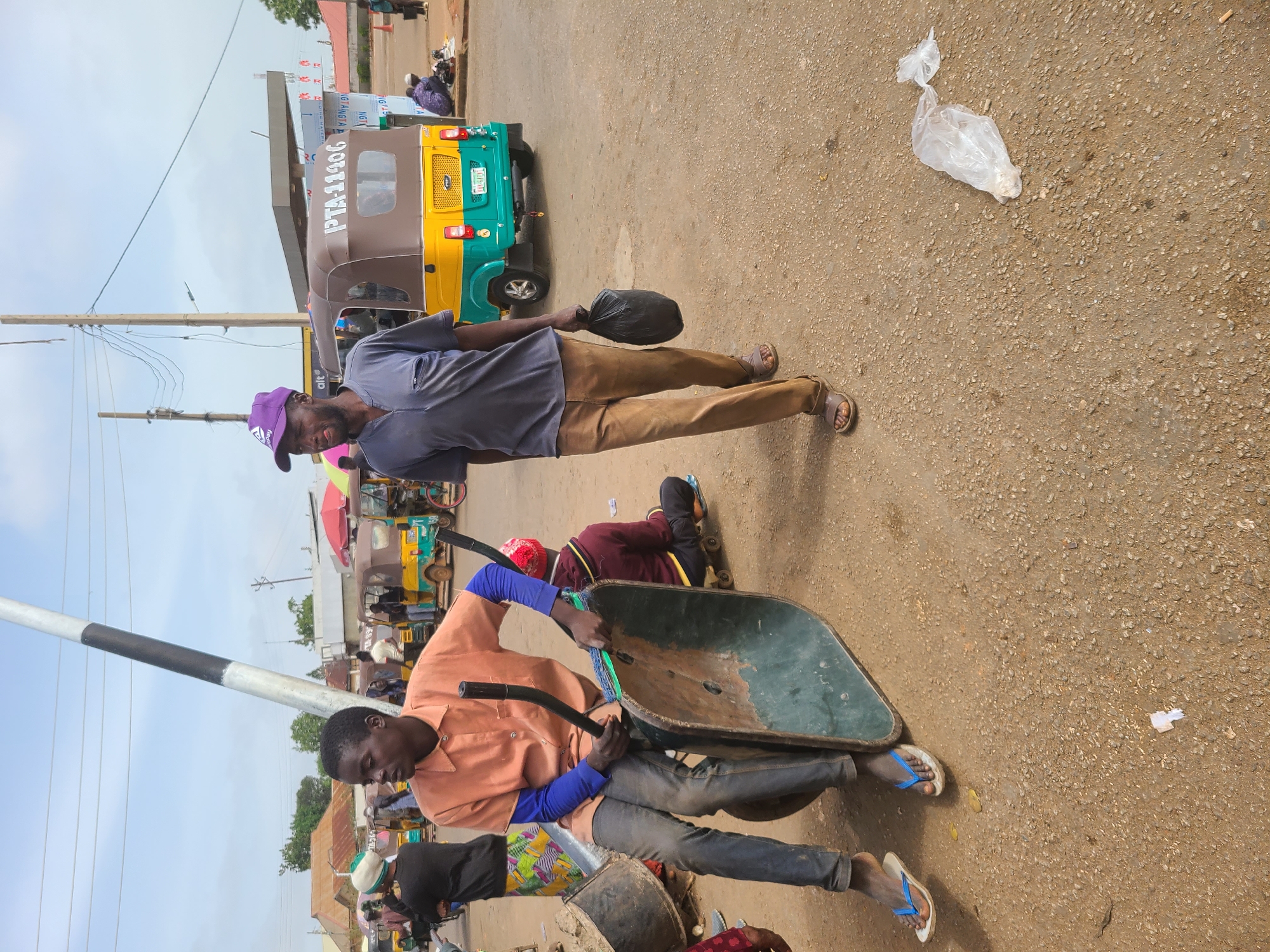 Two men are on a dirt road, one with a wheelbarrow. The other man carries a black bag. There are yellow and green auto rickshaws in the background. The location is a city, Unknown, Unknown.
