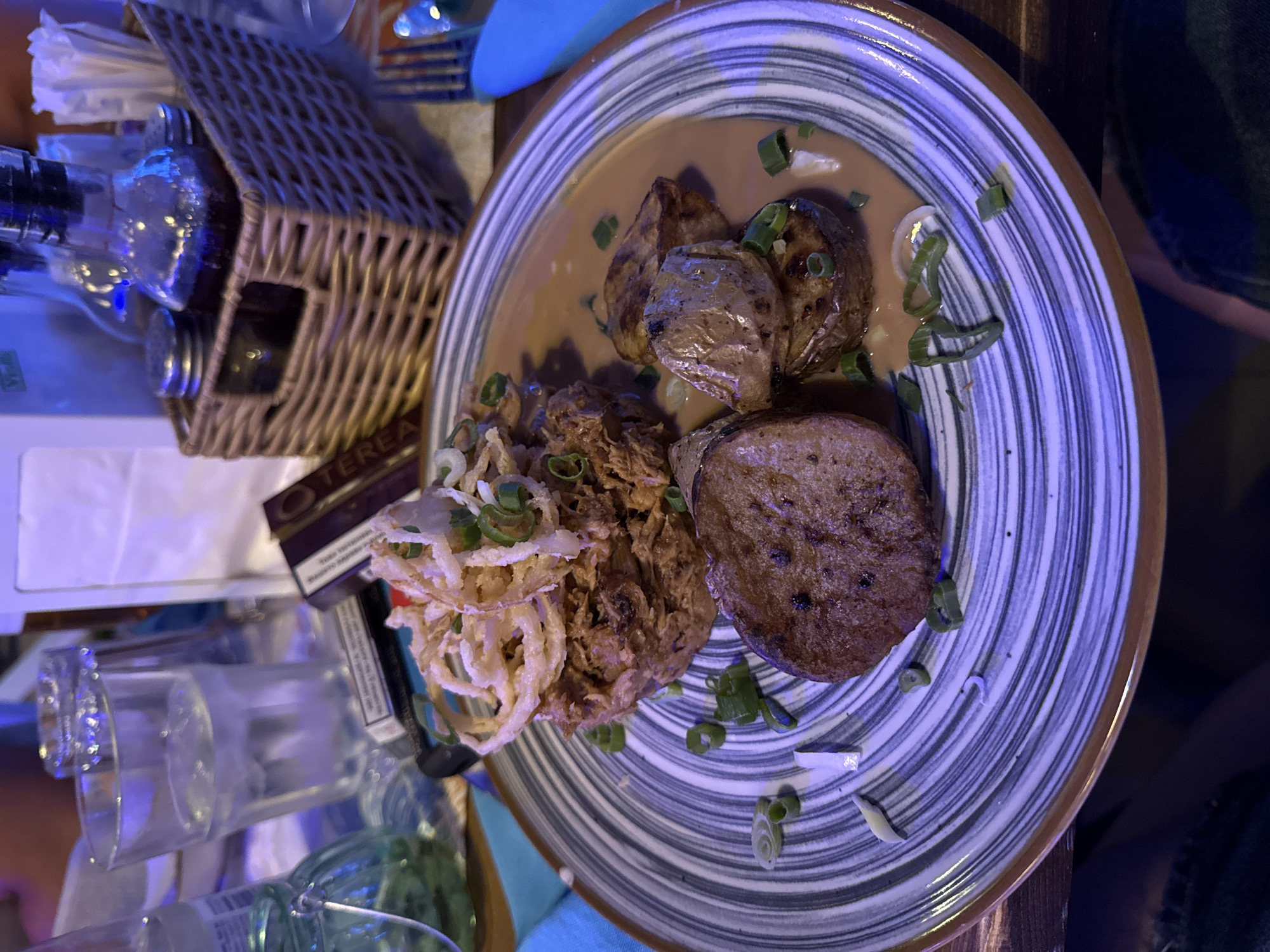 The image depicts a close-up shot of a plate of food, likely taken in a restaurant. The plate contains potatoes in a brown sauce with green onions, fried onion rings, and shredded meat, potentially chicken or pork. The plate has a spiral pattern with a brown rim. In the background, on the left side of the image, there is a small wicker basket, a bottle, and some glasses. A pack of Terea sticks is also visible next to the plate. The setting appears to be a restaurant table. The food presentation is somewhat rustic. While the image gives the impression of a dining experience, it is impossible to concretely pinpoint the location within Plovdiv, Bulgaria, from the information provided in the image.