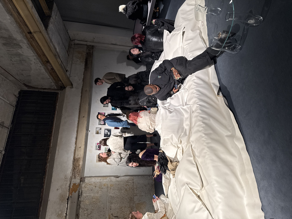 The image depicts an interior scene of a social gathering or exhibition space in Paris, France. The environment features aged concrete walls with visible texture and wear, an exposed structural beam, and a dark, possibly grated, overhead ventilation or window structure near the ceiling. The floor is covered with a dark blue or gray carpet. Along a main wall, several framed photographs or artworks are displayed.
In the foreground and midground, a large arrangement of oversized white upholstered cushions or mattresses forms a central seating area. A beige garment and some black fabric are placed on this white structure. Adjacent to it, a clear glass coffee table with organic-shaped legs holds two clear glass vessels, one containing a yellowish liquid.
Approximately 15 to 20 individuals are present. Several people are standing in small groups near the wall display, engaged in conversation. Other individuals are seated or lounging on the white cushioned structure or on low benches/the floor. A man wearing a dark suit with a light-colored embellishment on his lapel is seated on the edge of the white structure in the foreground, looking at a smartphone while holding a clear glass containing a light-colored drink. A woman with long dark hair, wearing a purple velvet top, is seated on the floor next to the white structure. Another individual with reddish-pink hair is seated further back on the floor. Interactions primarily involve conversational groupings and individual observation of personal devices or the surroundings.