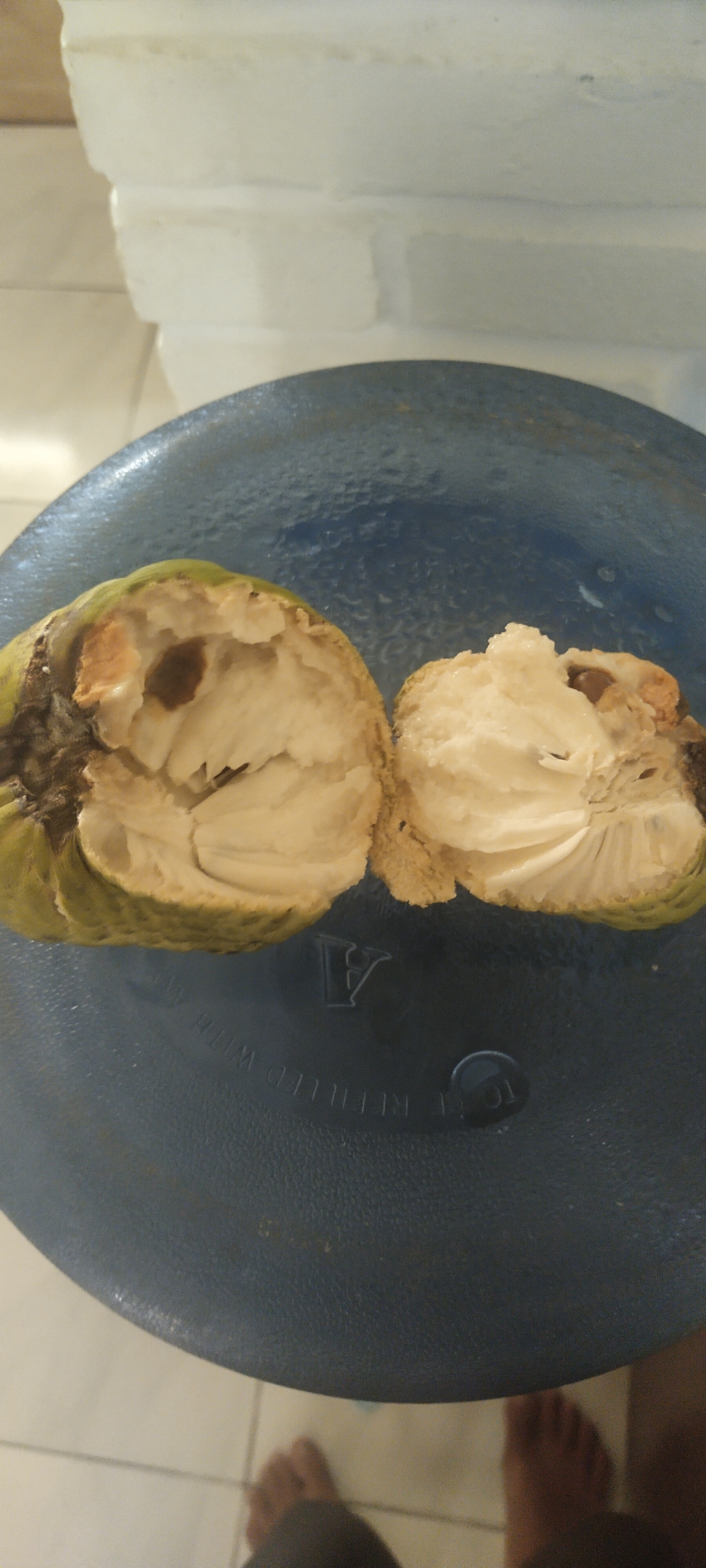 In Pasirhuni, Indonesia, a cut soursop rests on a dark blue circular surface, possibly a container lid. The fruit, split lengthwise, reveals a creamy white interior with visible dark seeds. The outer skin is green with some brown, slightly damaged areas. In the background, a white, brick-like structure is visible. The scene is illuminated by artificial light, casting faint shadows. Two bare feet are visible at the bottom of the image.