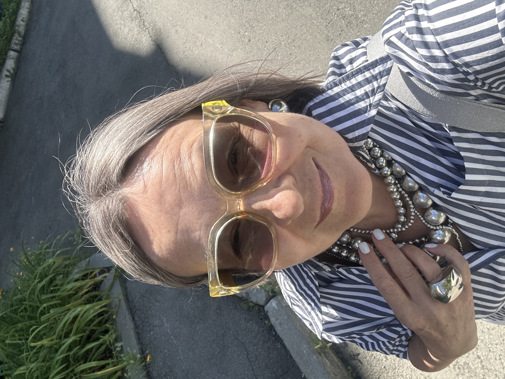 An outdoor, eye-level, medium shot captures a woman from the chest up. She has short, gray hair and is wearing large, translucent yellow-rimmed sunglasses. Her attire includes a blue and white striped button-up shirt, a chunky silver bead necklace, and a large silver ring on her left hand. A grey strap from a bag is visible over her right shoulder. She is standing on a paved surface, possibly a sidewalk or street, with a strip of green foliage and yellow flowers to her left. The background shows more paved area and a shadow cast by an unseen object. The lighting suggests it is daytime with bright sunlight.