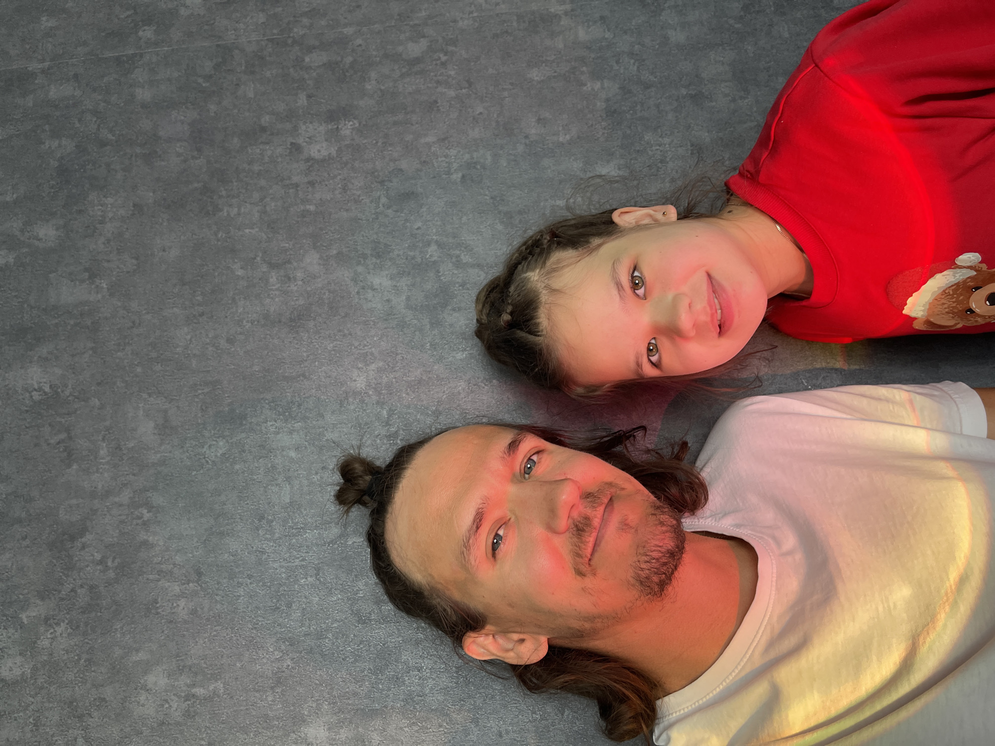 Two individuals, an adult male and a young girl, are lying on their backs on a dark gray, textured surface, likely a floor. The image is taken from a top-down perspective, with both subjects looking up towards the camera.
The adult male, positioned closer to the bottom of the frame, has long, dark hair pulled into a small bun at the back of his head and stubble/a beard. He is wearing a white t-shirt. A distinct, colorful light reflection, similar to a rainbow or prism effect, falls across his face and the upper part of his t-shirt, illuminating it with hues of red, orange, and yellow.
The young girl, situated above and to the right of the male, has dark hair styled in braids. She is wearing a bright red long-sleeved top featuring a graphic of a teddy bear wearing a Santa hat. A similar colorful light reflection is visible across her red top, and her face is also softly illuminated with warm tones. She has a subtle smile on her face. Both individuals appear relaxed and calm, posing for the photograph. The background is uniform, providing no additional environmental context beyond the floor texture. There is no visible text.