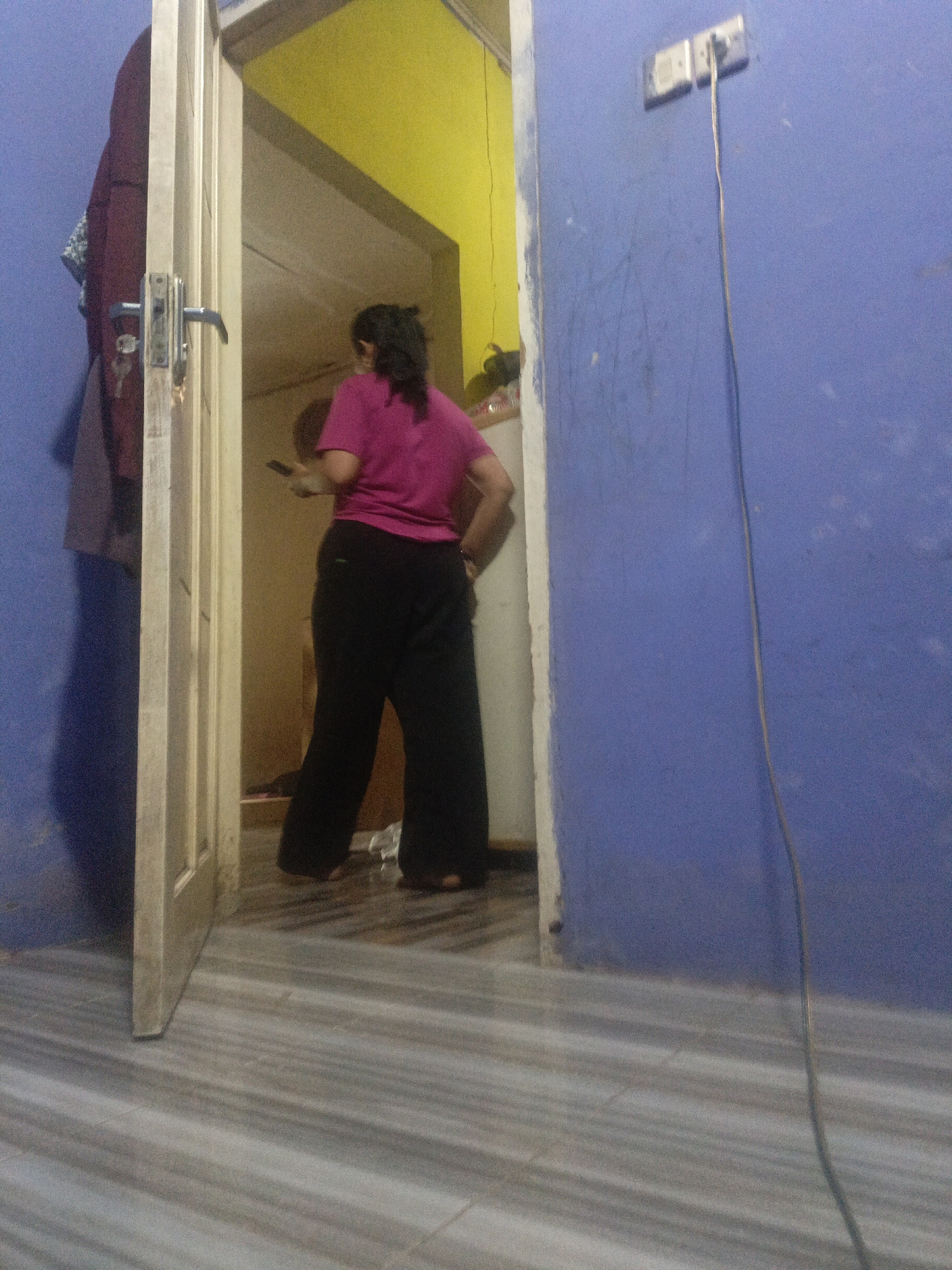 A person wearing a pink t-shirt and black pants stands inside a doorway, facing away from the camera and holding a black object, possibly a phone, in their right hand. The doorway opens into a room with yellow walls. To the left of the doorway, a maroon coat hangs on a hook attached to a light-colored door frame. A blue wall with an electrical outlet and a dangling wire is visible to the right of the doorway. The floor is tiled with a gray and white striped pattern. The scene appears to be in a residential interior, potentially in Lubangbuaya, Indonesia.