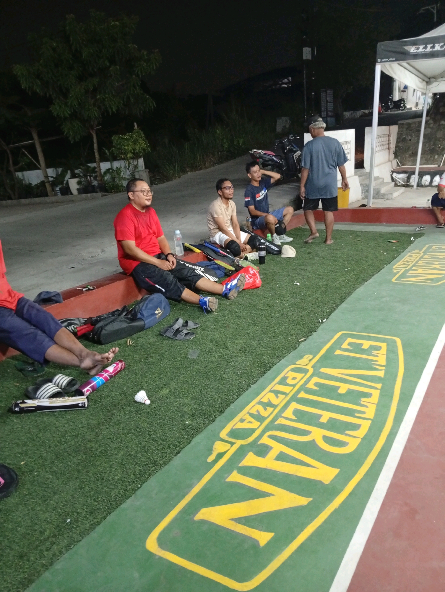 The image depicts a group of men gathered outdoors at dusk, on what appears to be a sports court with artificial turf and a red adjacent surface. The background suggests a semi-urban or recreational setting in Caringin, Indonesia, with trees, buildings, and overhead lighting visible.
In the foreground, several men are seated on a low red border. One man in a red t-shirt and black shorts is looking to his left. Next to him, a man in a light brown t-shirt and white shorts is also seated. Further left, another man in a blue t-shirt and dark shorts is sitting with his arm behind his head. A fourth man, wearing a grey t-shirt and black shorts, is standing and facing away from the camera, appearing to be talking to the seated group. He is barefoot.
Scattered on the artificial turf are various objects, including badminton rackets and bags, a water bottle, slippers, and a shuttlecock. A large green banner with yellow lettering that reads "VETERAN PIZZA" is prominently displayed on the ground in the lower right quadrant of the image. The overall scene conveys a relaxed atmosphere, possibly after a game or during a break.