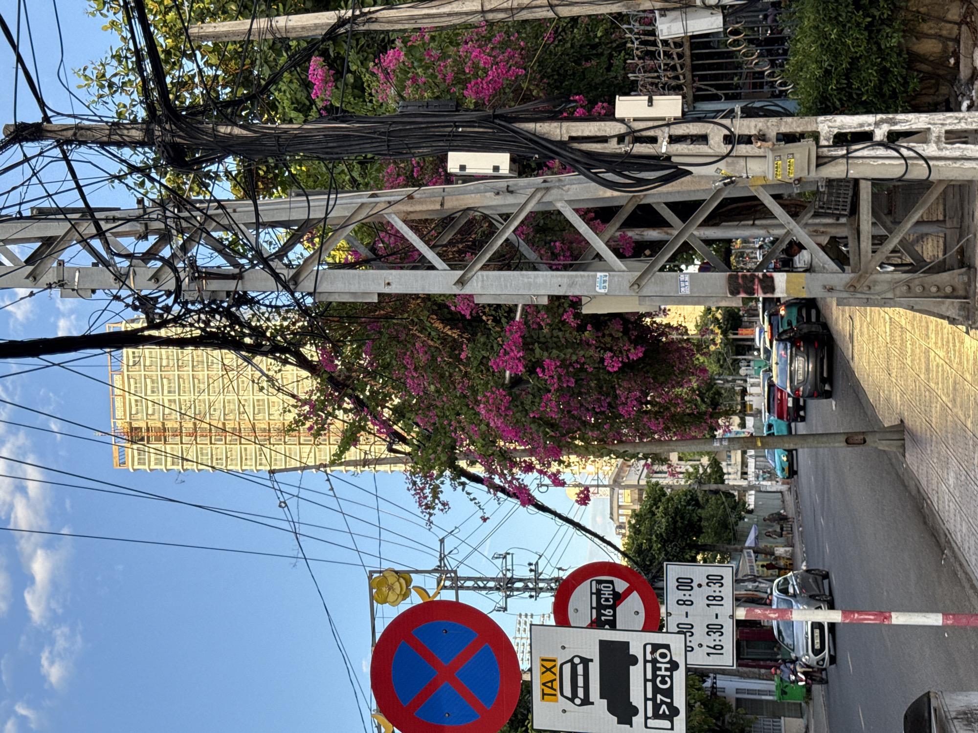 The image depicts a street scene in Nha Trang, Vietnam, characterized by a tangle of electrical wires overhead. In the foreground, a series of traffic signs are visible. A circular red and blue sign with a red "X" indicates no parking. Below this, a white sign displays "TAXI" at the top, followed by icons of a car and a truck, and the text ">7 CHO". Another circular red sign shows "16 CHO" within a white circle. A rectangular sign underneath lists time restrictions: "6:30 - 8:00" and "16:30 - 18:30". A street pole with red and white stripes stands next to these signs. The street itself is paved, with cars and a motorcycle visible on it. To the right, a sidewalk is lined with a metal fence and lush green foliage with pink flowers. In the background, a tall, multi-story building under construction, with exposed concrete and scaffolding, dominates the skyline, contrasting with the clear blue sky.