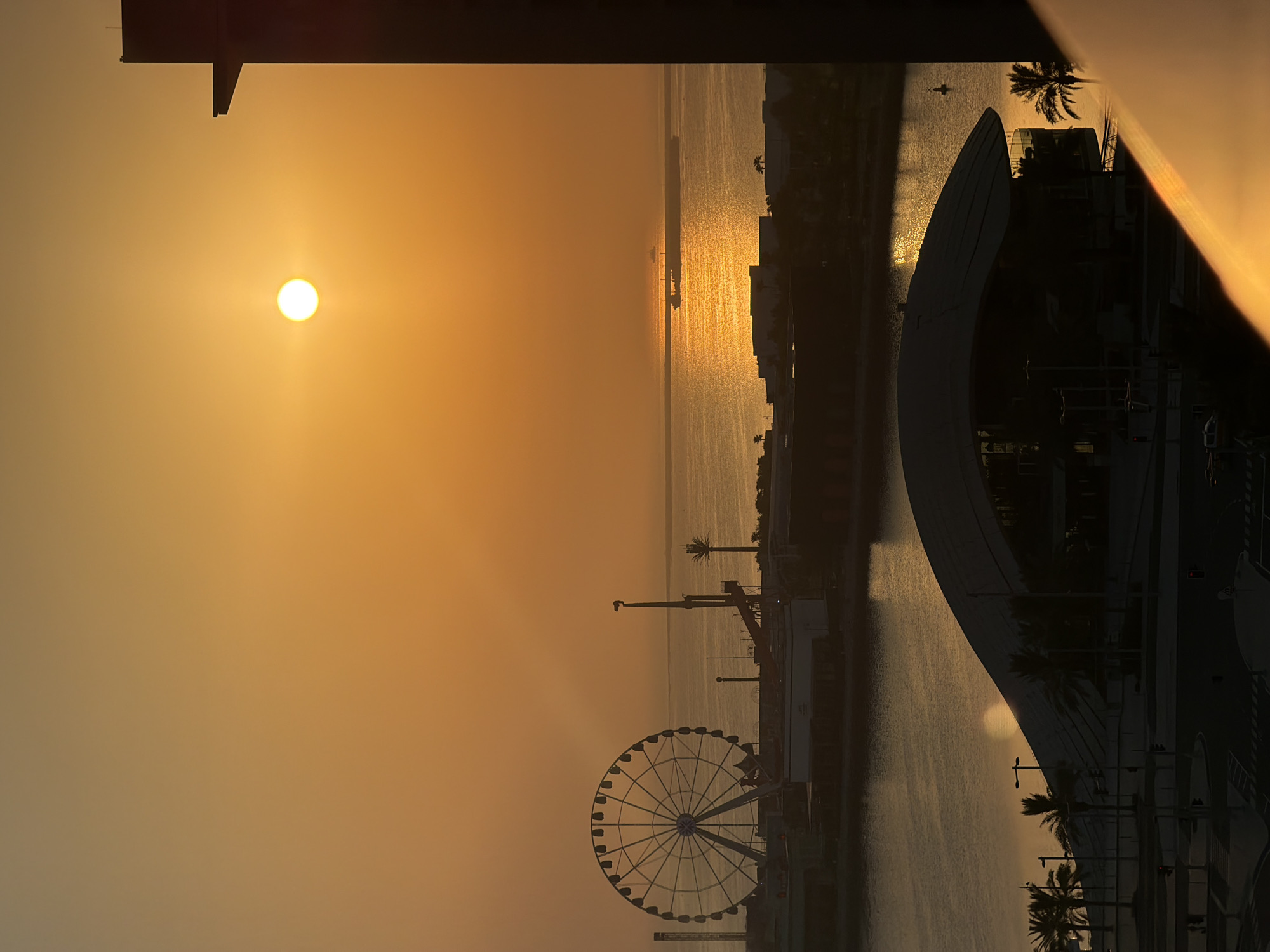 An aerial, high-angle, outdoor, vertical, long shot captures a sunset over a body of water and a cityscape. The sun is a bright orange orb in the hazy, orange sky, casting a shimmering golden reflection on the water. In the mid-ground, a Ferris wheel stands silhouetted against the sky, with a modern, curved building in the foreground. Palm trees are scattered throughout the scene. Several ships are visible on the horizon. The overall mood is serene and warm, bathed in the glow of the setting sun.