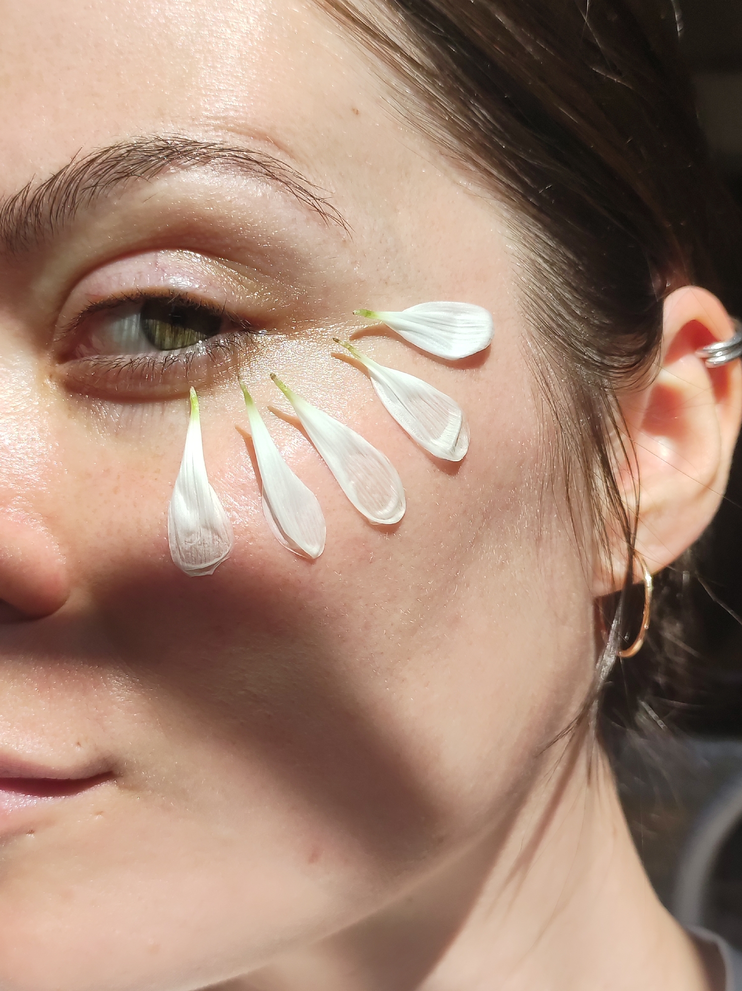 A close-up image shows a woman's face in sunlight, with several white flower petals arranged under her eye, appearing like stylized tears. The woman has fair skin, brown hair pulled back, and green eyes. She wears two earrings in one ear, a hoop and a stud. The flower petals are small, with a slight green hue at the base where they would connect to the flower. The background is out of focus, but appears to be outdoors. There is no evidence that this image was taken in Фоки, Russia.