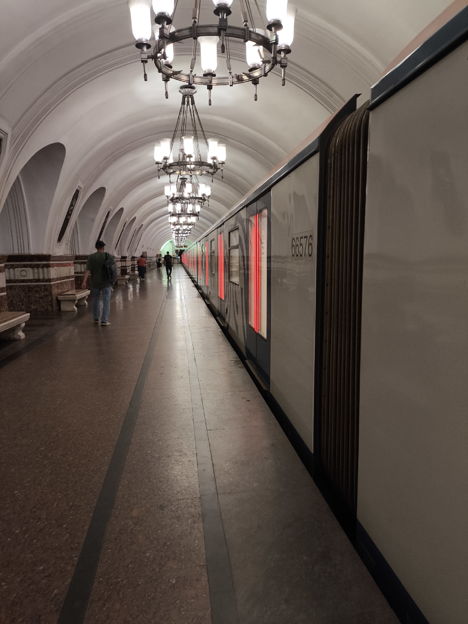 An interior shot captures a subway platform in an unknown location. The platform is lined with a train on the right, its windows illuminated by red light. A man with a backpack walks away from the camera down the platform, with other figures visible in the distance. The arched ceiling is adorned with ornate chandeliers, and the walls are decorated with marble. The floor of the platform is a speckled red tile.