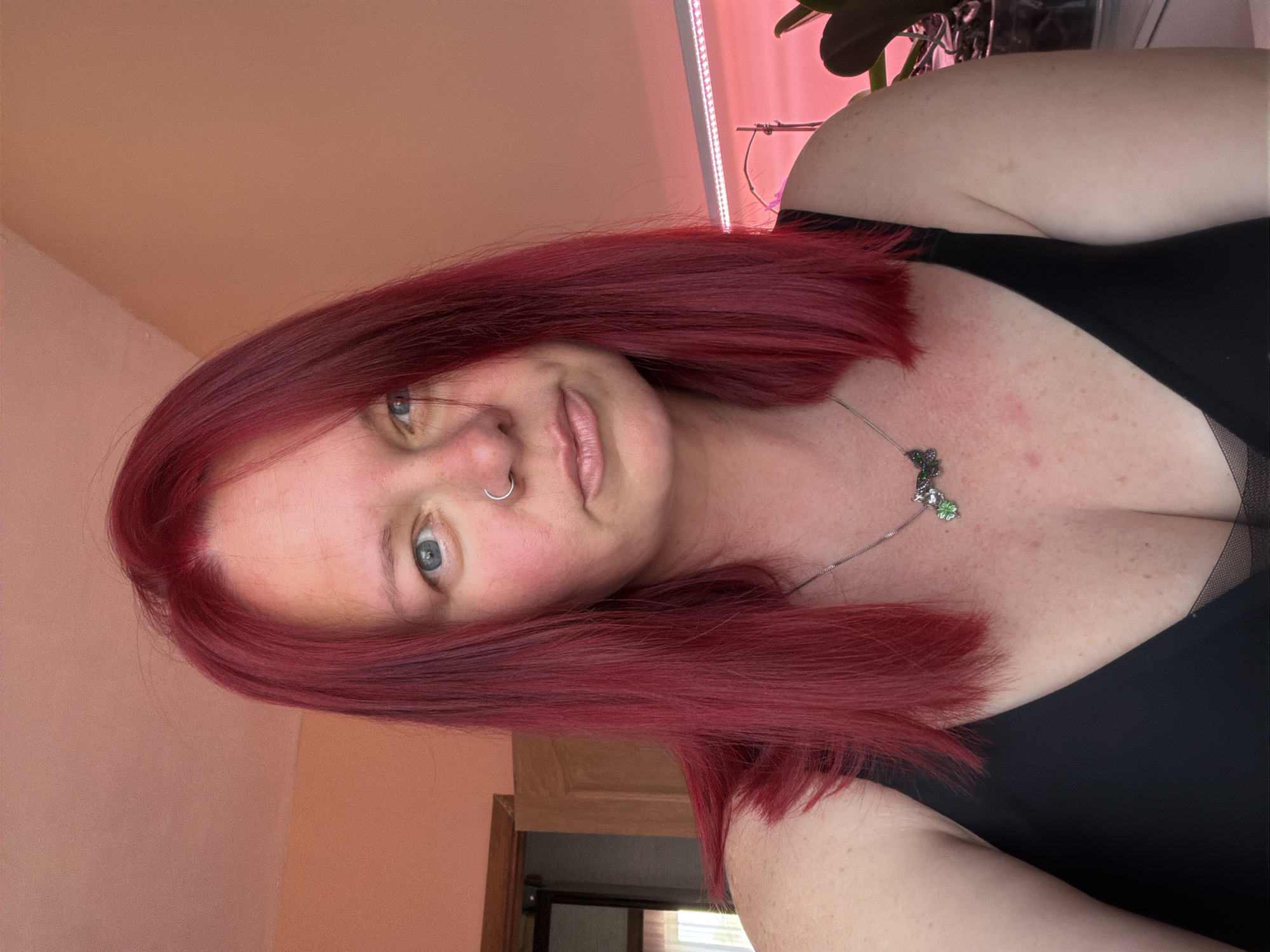 A person with red hair, a nose ring, and wearing a black top is in a room. The room has a peach-colored wall, a wooden door and frame, and a bright light fixture. A plant is visible in the background. The person is wearing a necklace with a clover charm. The image does not provide information to conclude the location as the city of Chaikovsky, Russia.
