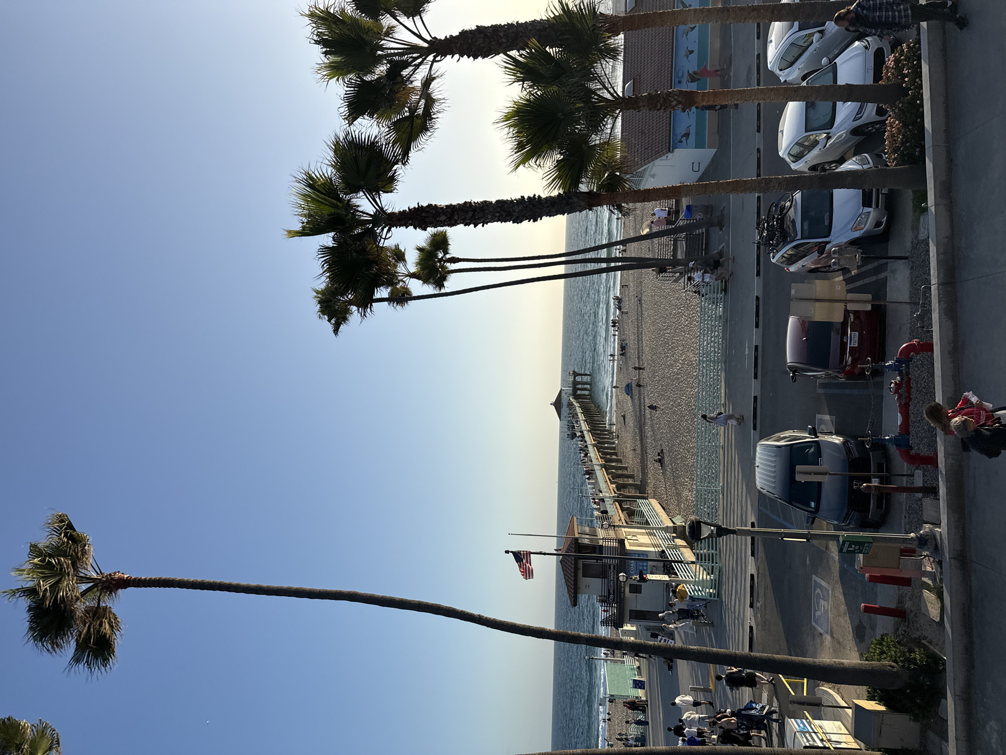 The image depicts a view of Manhattan Beach Pier in Manhattan Beach, California, United States of America. The pier extends into the ocean, with people visible walking on it and along the sandy beach. A lifeguard station with an American flag is situated near the base of the pier. Several palm trees line the foreground and the area near the beach, characteristic of the Southern California coastline. In the background, the ocean meets the horizon under a clear blue sky, with the sun casting a warm glow. In the foreground, there is a parking lot with several cars, including a black SUV, a red sedan, and a silver SUV with bicycles mounted on its roof rack. People are seen walking in the parking lot and on the sidewalk adjacent to the beach. The scene captures a lively atmosphere with people enjoying the beach and pier.