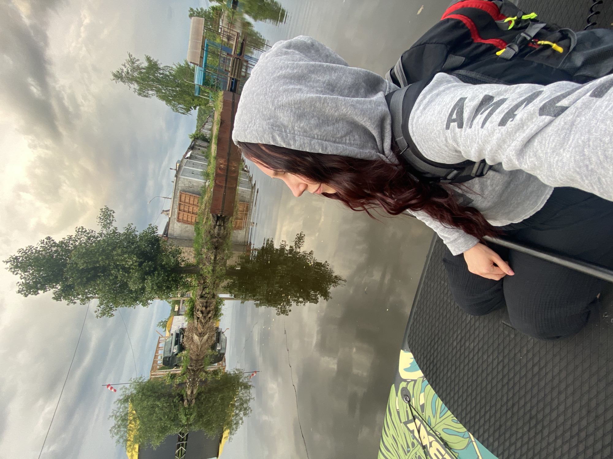 An overhead-angled shot shows a person on a paddleboard on a calm body of water, likely a river or canal, in Chaikovsky, Russia. The image is oriented with the person on the right, facing towards the left, and the shoreline on the far left.
The main subject is a person, likely female, with long, dark reddish-brown hair, wearing a grey hooded sweatshirt and dark pants. They are kneeling or crouching on the black, textured grip pad of the paddleboard. A black backpack with red and yellow accents is on their back. Their right hand is near a black paddle shaft. On their right sleeve, the letters "AMA" are visible in dark grey text. The paddleboard features a vibrant green, blue, and yellow tropical leaf pattern on its front section.
The water, which takes up a significant portion of the image, is still and reflects the overcast sky, trees, and distant buildings. Along the distant shoreline, there are several structures: some appear to be older, industrial-type buildings, possibly docks or workshops, with one having prominent wooden doors. A long, dark, possibly metallic structure runs along the water's edge. Green trees line parts of the bank, and their reflections are visible in the water. Further down the shore, a truck is partially visible, and a red and white striped windsock stands against the cloudy sky. The sky is largely overcast, suggesting diffused light, possibly from an early morning or late afternoon, or a cloudy day. The overall atmosphere appears calm and contemplative.