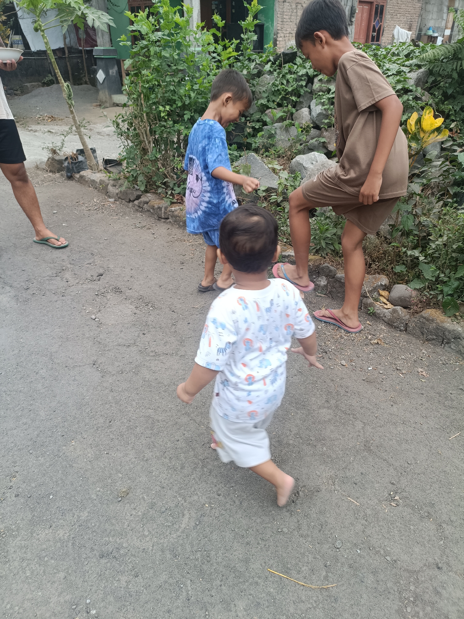 In Sumberadi, Indonesia, three young boys are in motion on a paved surface. The boy in the foreground, wearing a white t-shirt with a playful, patterned design and light-colored shorts, is walking away from the camera with his arms outstretched. To his left, a boy in a blue tie-dye t-shirt and dark shorts appears to be looking down. Further to the right, a boy in a brown t-shirt and shorts is captured mid-stride, with one leg lifted. The environment is characterized by a rough, textured ground, bordered by a collection of rocks and lush green foliage. In the background, a simple building with a red door is visible.