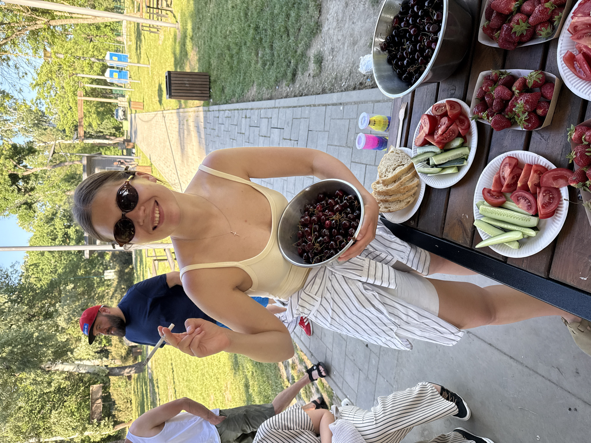 The image shows a gathering of people outdoors, potentially in a park, within the Cukarica Urban Municipality, Serbia. A fair-skinned woman in sunglasses and a light-colored tank top is holding a bowl of cherries. On a wooden table nearby are bowls and plates filled with cherries, strawberries, sliced tomatoes, cucumbers, and bread. The scene suggests a picnic or outdoor meal. A man in a blue shirt and red cap is visible in the background, holding what appears to be a cigarette. Additional people are seated or standing around the table, though they are partially obscured. The environment consists of grass, trees, a paved path, and park benches.