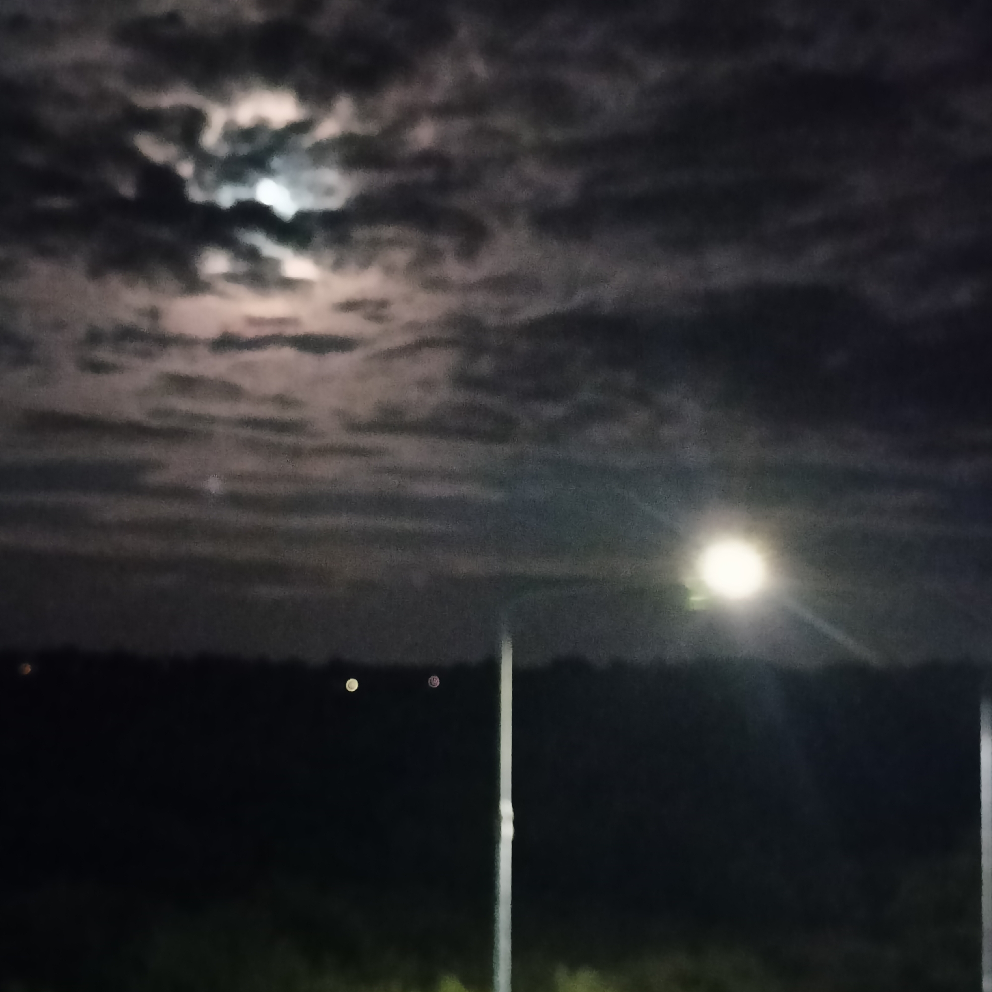 The image depicts a nighttime scene under a cloudy sky. A bright, full moon is partially obscured by dark clouds in the upper left portion of the frame. In the mid-ground, a bright street light illuminates the scene from the right, casting a distinct glow. A tall, slender object, possibly a lamppost, is visible in the lower right quadrant, its light extending upwards. The foreground is dark and appears to be an outdoor area with some vegetation, suggested by a faint green hue. In the far distance, a dark line indicates a horizon with a few small, indistinct lights, suggesting a populated area or a distant town. The overall impression is one of a quiet, possibly rural or suburban, evening in Limerick, Ireland.