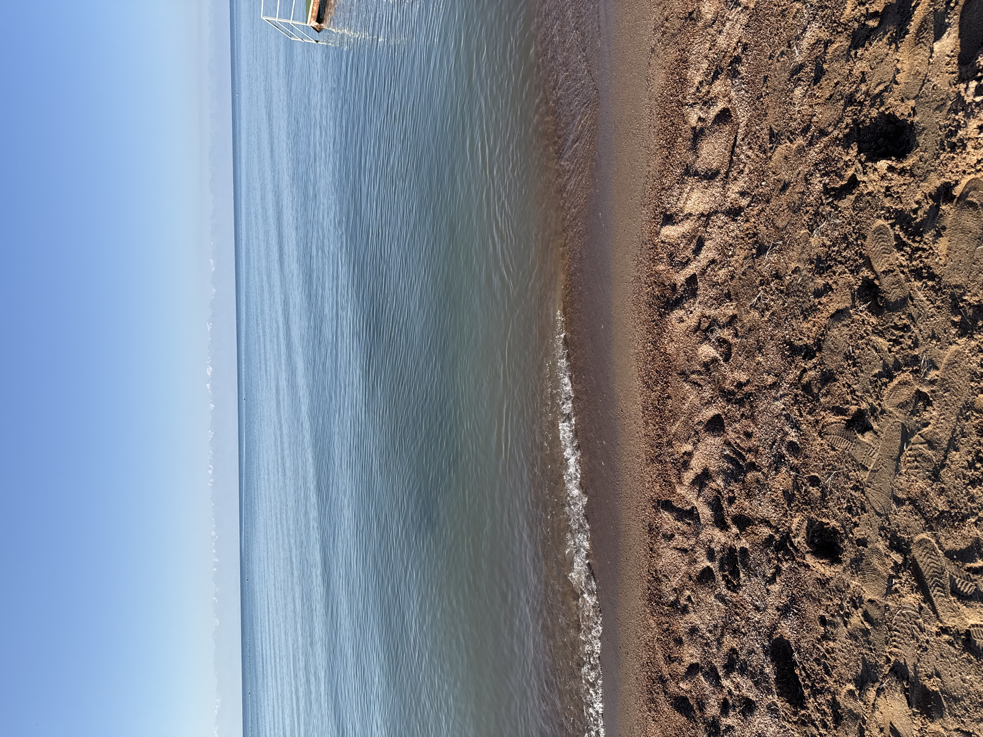 An outdoor, eye-level, horizontal shot shows a serene lake scene under a clear blue sky in Sary-Oy, Kyrgyzstan. The foreground is dominated by a sandy beach with numerous footprints and tire tracks imprinted in the sand. The sand appears dry and coarse.
The middle ground features the calm, clear water of a lake, with gentle waves lapping at the shore. The water transitions from a lighter, translucent hue near the beach to a deeper, more opaque blue further out. In the distance, a hazy line of snow-capped mountains stretches across the horizon, under a pale blue sky that gradually darkens to a deeper blue at the zenith. On the right side of the image, a white railing attached to a wooden platform extends slightly into the water, suggesting a pier or dock. The overall atmosphere is peaceful and tranquil.