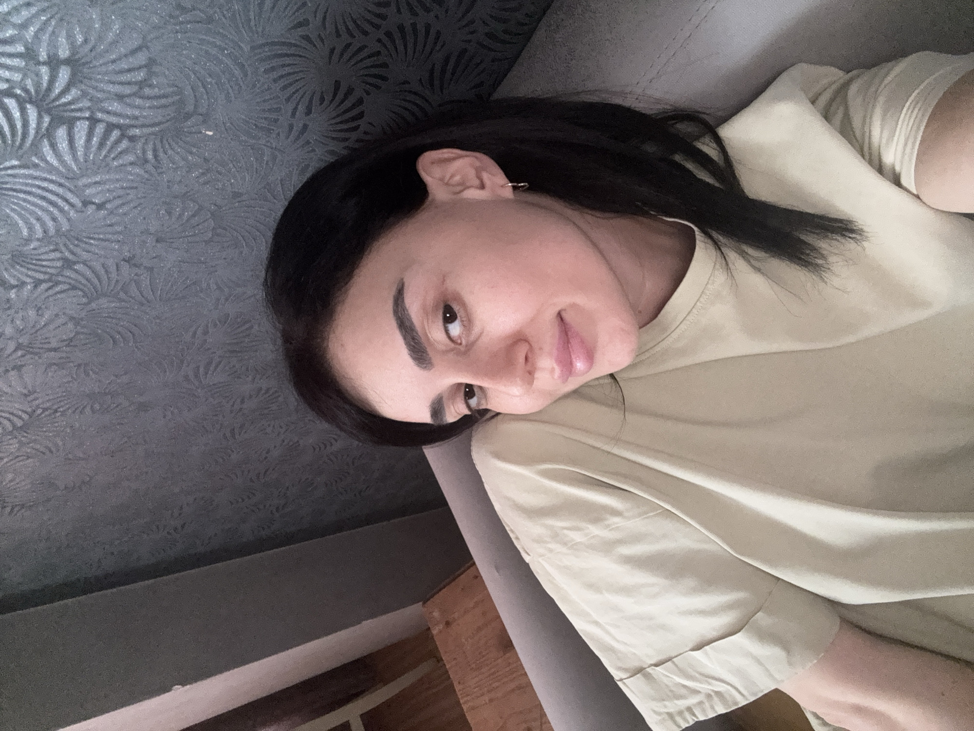 A person with dark hair is lying on a grey couch, with their head resting on a grey cushion. They are wearing a light tan-colored, long-sleeved shirt. The background features a patterned grey wall. The location is indoors, details about the location in the city of Tbilisi, Georgia cannot be determined from the image.
