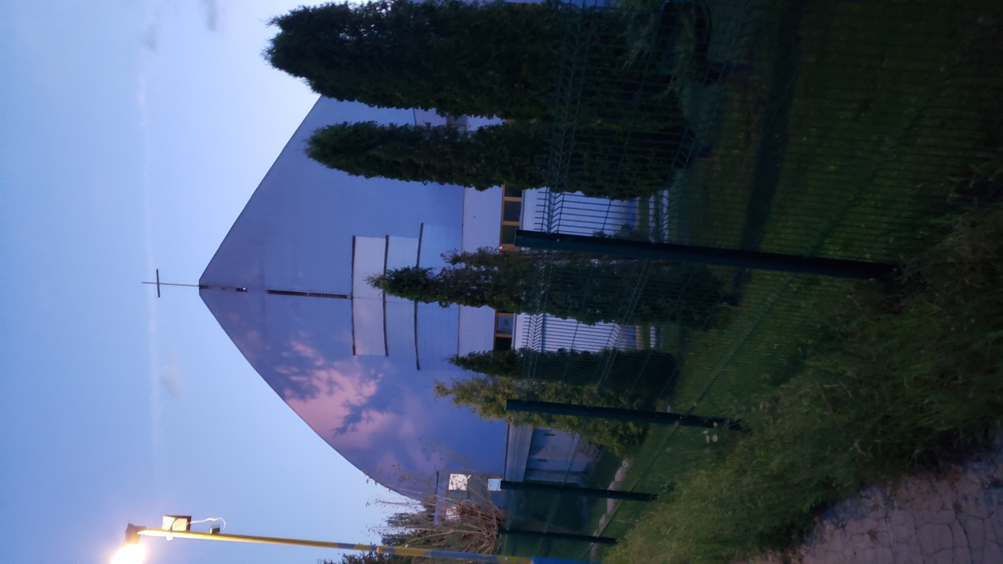 The image captures the exterior of a modern church building with a distinctive, rounded roofline, located in Prešov, Slovakia. A cross is prominently positioned at the apex of the roof. The church's facade is a muted blue, with visible horizontal architectural elements and windows. In front of the building, a black metal fence encloses an area with several tall, slender evergreen trees. To the left of the church, a streetlamp with a yellow pole and a light fixture casts a glow against the twilight sky. The foreground shows a paved walkway and a grassy verge bordering the fence. The sky is a pale blue, transitioning into darker hues of twilight, with faint clouds reflecting pinkish hues from the setting sun. The scene appears to be a still, quiet evening in a suburban or semi-urban setting.