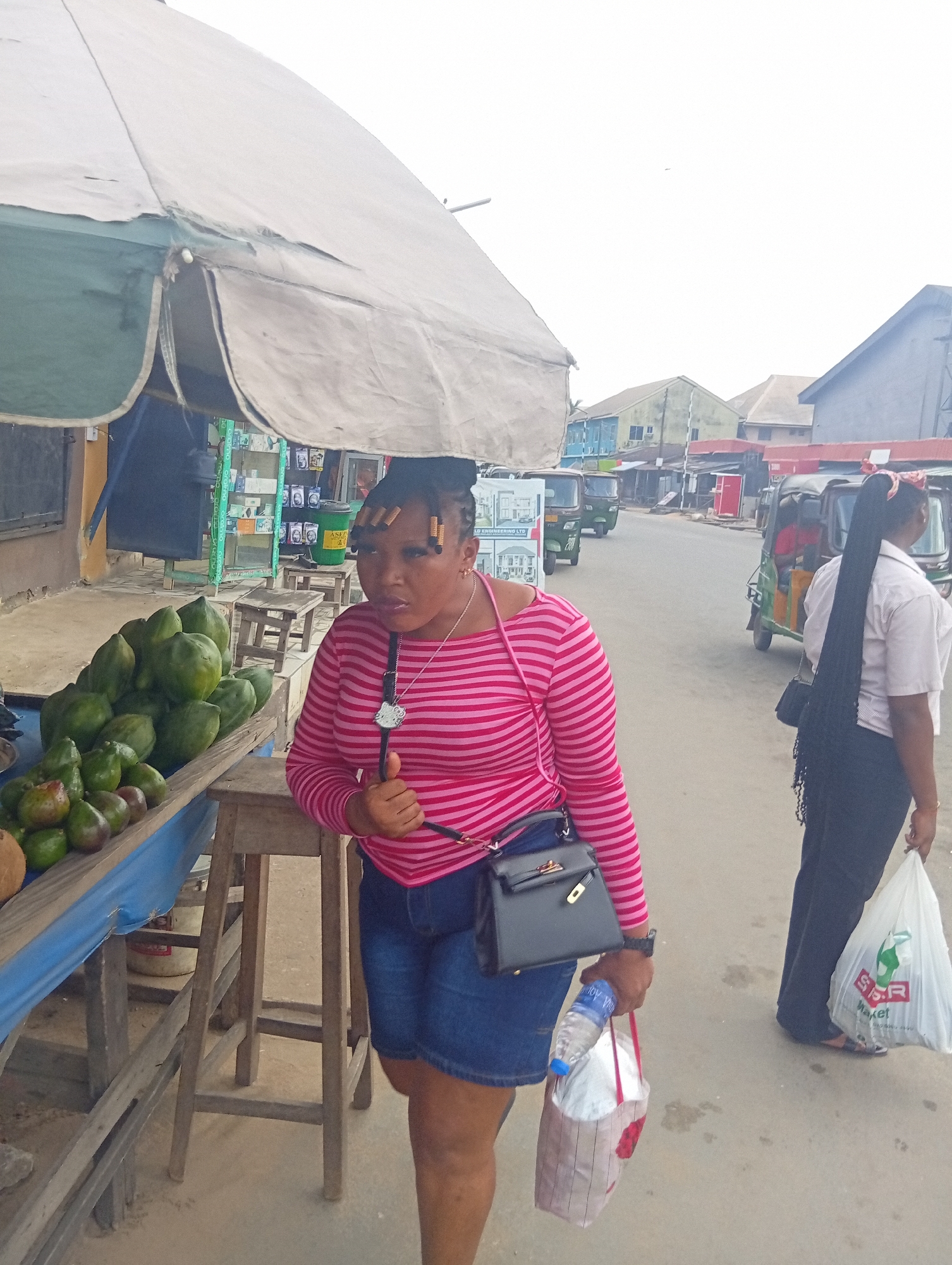 The image captures a bustling street scene, likely a market area, in Aba, Nigeria. The primary subject is a young woman standing at the edge of a roadside stall, her left arm resting on a wooden stool. She wears a pink and white striped long-sleeve top, denim shorts, and a black handbag with a gold clasp. A silver necklace with a Hello Kitty pendant hangs around her neck. Her hair is styled with small, cylindrical, light-colored hair accessories. She holds a plastic water bottle and a small patterned shopping bag in her right hand, her expression appearing somewhat serious or contemplative.
To her left, the stall displays a large pile of green, unripe papayas or avocados, along with other fruit like coconuts, on a wooden surface covered with a blue tarp. Behind the stall, additional wooden stools and shelves with various products are visible, including a green bucket with the text "ASEP".
In the background, a dirt road stretches into the distance, lined with various buildings, some appearing residential and others possibly commercial or market stalls. Three-wheeled vehicles, commonly known as "keke Napeps" in Nigeria, are visible on the road, with one moving away from the viewer. Another woman, seen from behind, walks away on the right side of the frame. She wears a light-colored shirt and dark trousers, and her very long, dark braided hair extends down her back. She carries a white shopping bag with "SPAR Market" printed in red and green text. The sky is overcast, suggesting either an early morning, late afternoon, or a generally cloudy day. The overall impression is that of everyday life in a lively urban or semi-urban Nigerian environment.