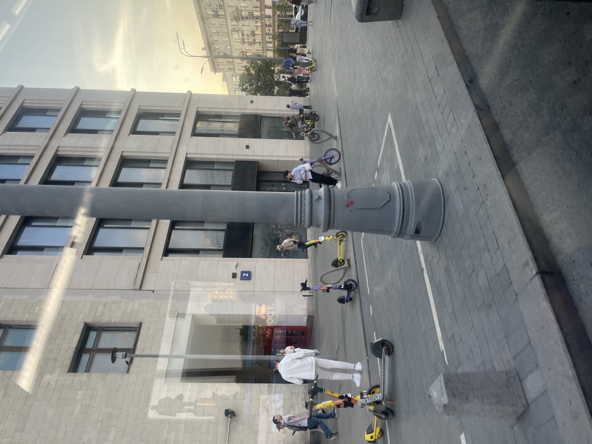 The image shows a street scene in Moscow, Russia, with a multi-story building on the left and a paved sidewalk on the right. People are visible on the sidewalk, some riding electric scooters, and others standing or walking. Several scooters are parked in designated areas marked with white lines. A gray lamppost stands in the foreground. The building has numerous windows and a sign with the number "2" is visible. A red kiosk or booth is also noticeable in the reflection of the building. The lighting suggests it is either early morning or late afternoon.
