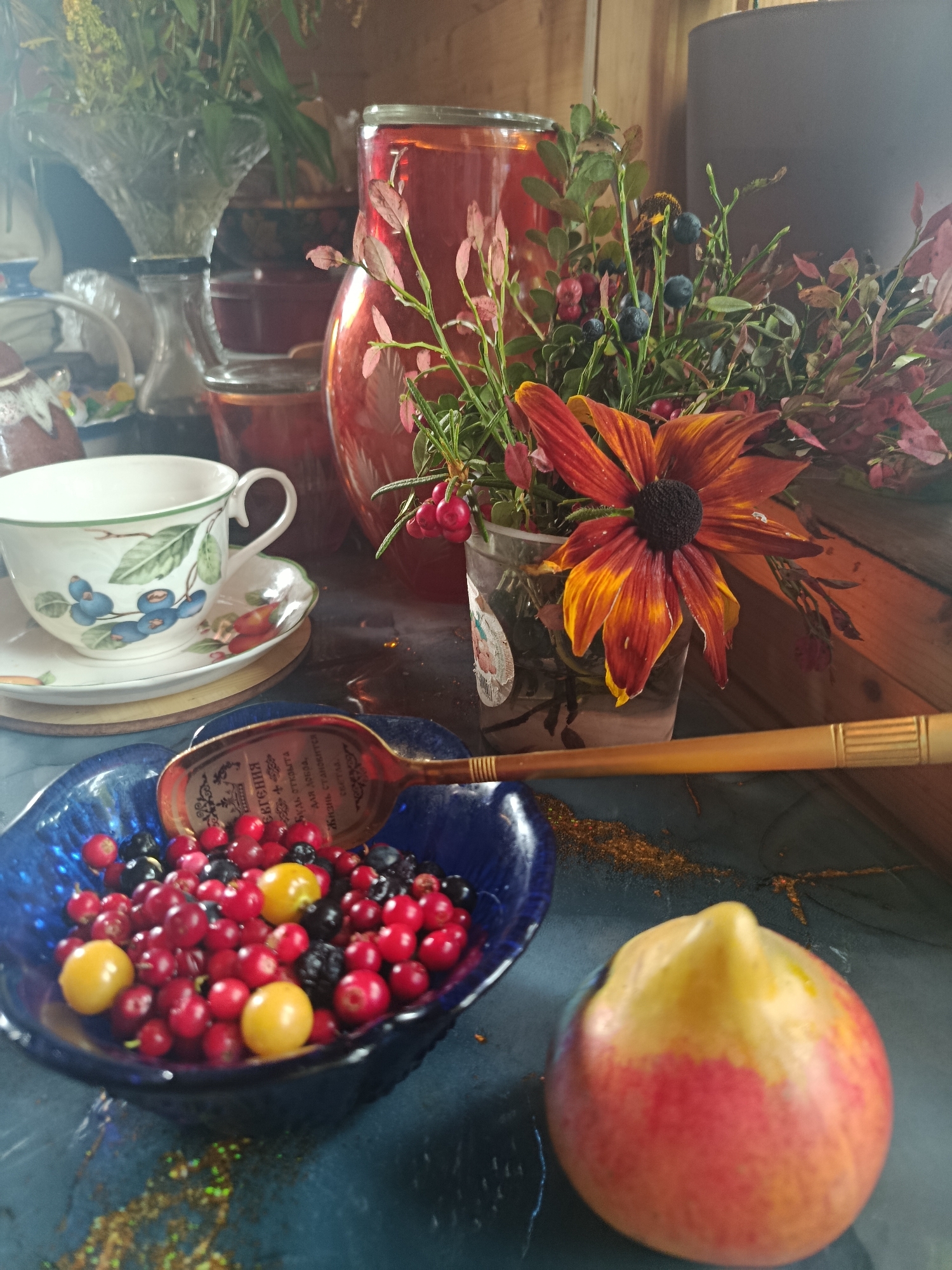 A still life arrangement is presented, featuring a blue bowl filled with assorted berries, including cranberries, blueberries, and gooseberries. A wooden spoon with an ornate inscription rests on the bowl. To the left, a white teacup with a blue floral pattern sits on a saucer. In the background, a tall, clear glass pitcher holds a dark red liquid, and a bouquet of flowers and greenery, including a large orange and yellow daisy, is displayed in a transparent container. A single apple, showing red and yellow coloration, is positioned in the foreground to the right. The setting appears to be indoors, possibly on a table with a textured, dark surface. The lighting suggests a natural light source, casting soft shadows. The image is devoid of any discernible urban landscape or specific city identifiers.