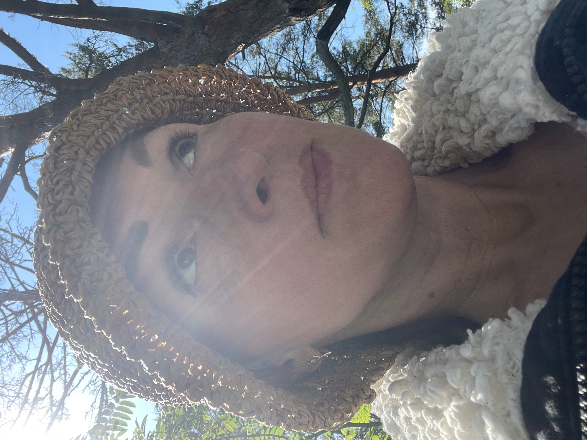 A woman wearing a straw hat and a white, fuzzy jacket looks upwards and to the right, bathed in bright sunlight filtering through tree branches. She has freckles on her face and is wearing a small, metallic earring. The background consists of a clear blue sky and the textured bark of a large tree. The scene is an outdoor setting, likely in Zelenaya Roshcha, Russia, given the presence of trees and the quality of the light.