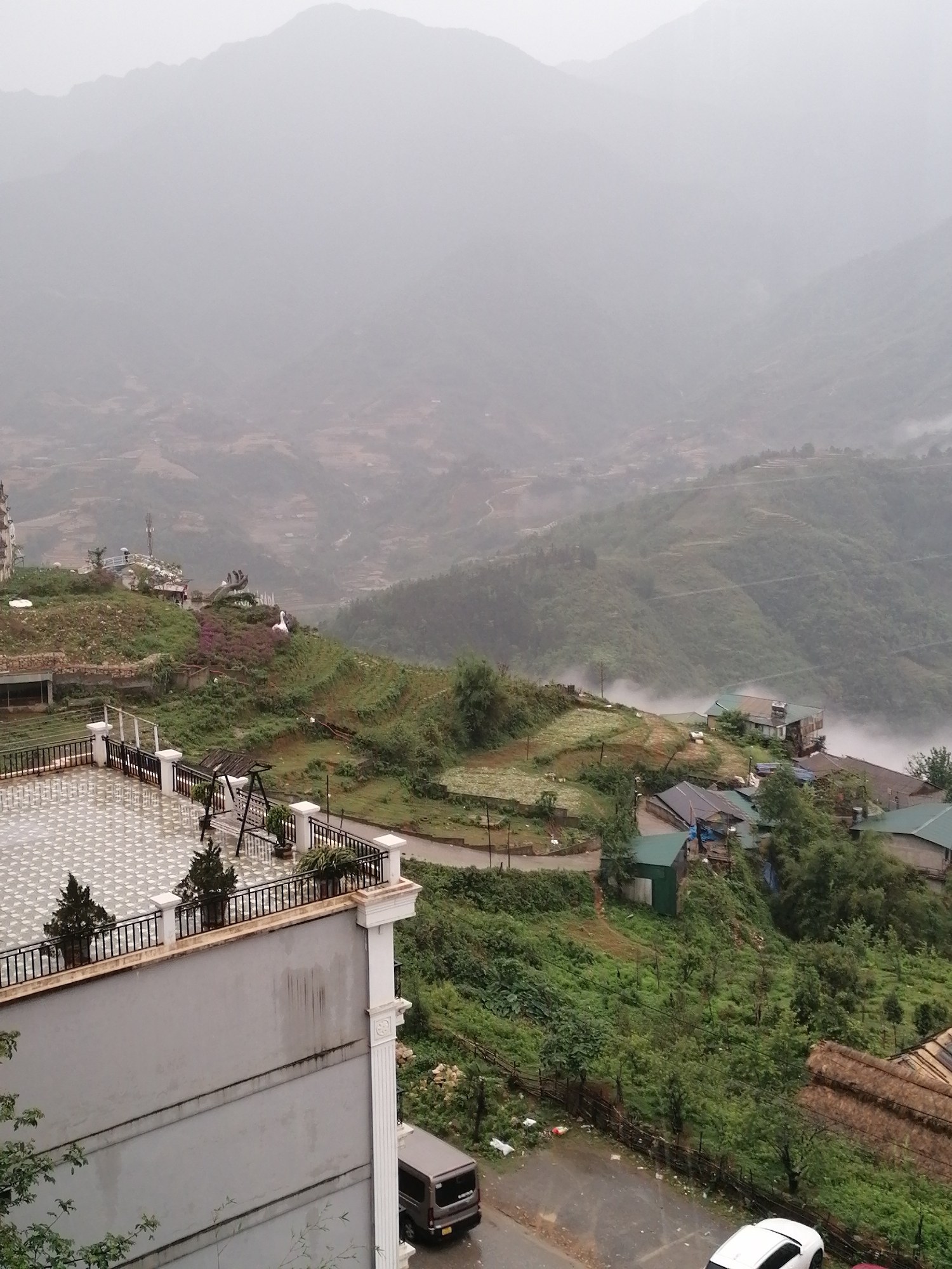 The image shows a hazy mountain landscape in Sa Pa Ward, Vietnam. In the foreground is a multi-story building with a tiled rooftop terrace that has potted plants and a swing set. Below the building are several parked cars. The middle ground is a green, hilly area with scattered buildings, gardens, and a winding road partially obscured by mist. The background reveals layers of mountains fading into a cloudy sky.