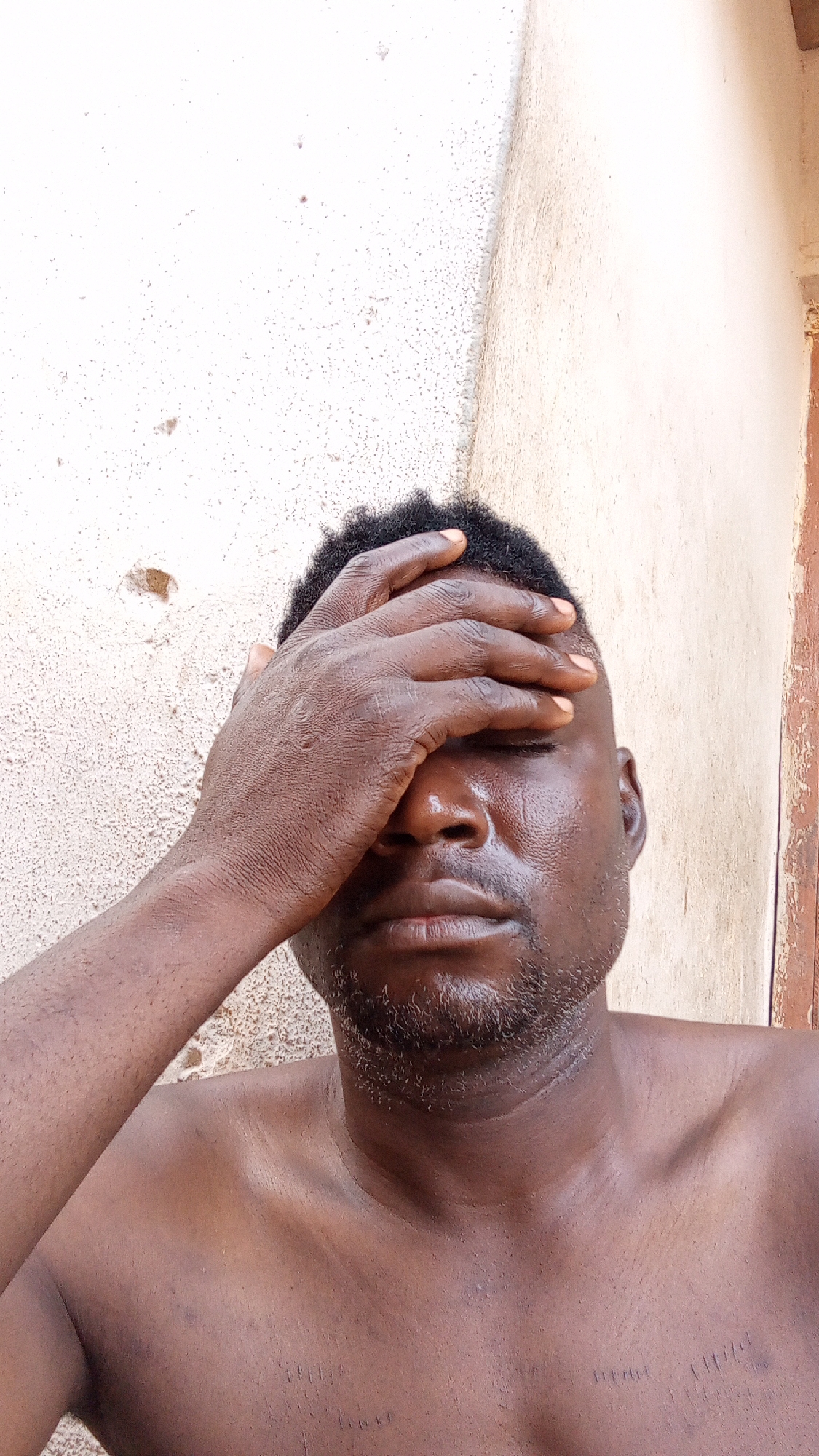 A man with dark skin and short, dark hair is shown from the chest up, with his hand covering his forehead and eyes. He appears shirtless. The background is a textured, light-colored wall, with a darker, vertical line on the right edge of the frame, potentially indicating a doorway or edge of a building. The location is Unknown, Unknown.
