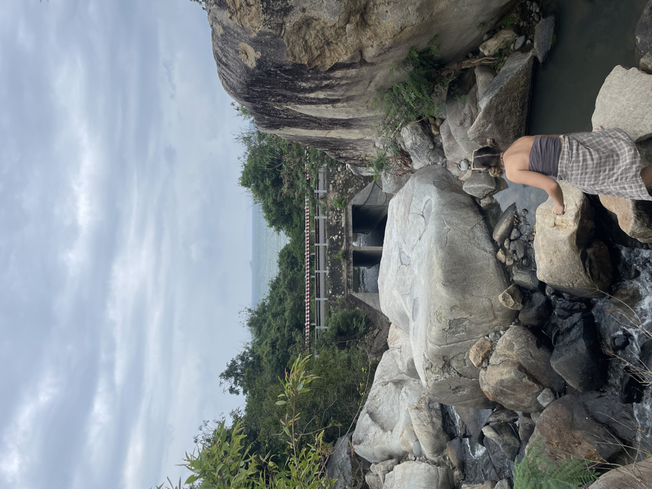 The image presents a natural landscape in Ninh Hòa, Vietnam, under an overcast sky. In the immediate foreground, a person, observed from behind, stands on a large, rounded, light-grey boulder. The individual wears a grey strapless top and a light-colored plaid shirt tied around their waist; their hair is styled in two buns. They face a stream bed densely filled with numerous large, smooth boulders of varying sizes and tones, through which water flows from left to right. In the midground, a concrete road bridge or culvert spans the stream, featuring at least two rectangular openings for water passage. A paved road, complete with a low guardrail marked with red and white stripes, runs across the top of this structure. Dense green tropical vegetation lines the banks of the stream and flanks the road. In the background, beyond the vegetation and road, the ocean is visible under the grey, cloudy sky, with a small, distant island discernible on the horizon.