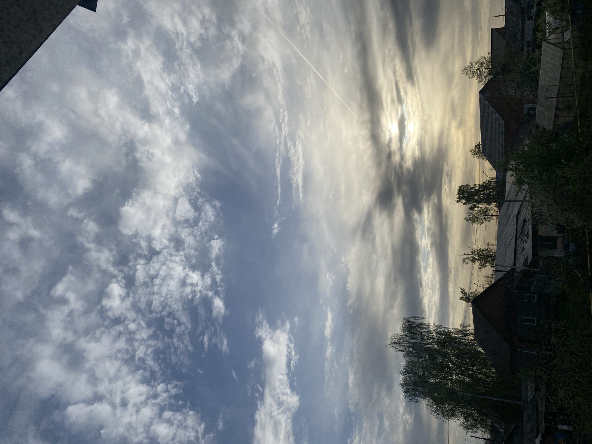 The image shows a daytime view from Chaikovsky, Russia. The sky dominates the frame, displaying a mix of cirrus and cumulus clouds with a contrail from an aircraft streaking across. The sun is partially obscured by a layer of clouds, casting a diffused light. In the lower right, the rooftops and tree canopies of houses are visible, appearing relatively dark due to the lighting conditions. The angle suggests the image was taken from a high vantage point.