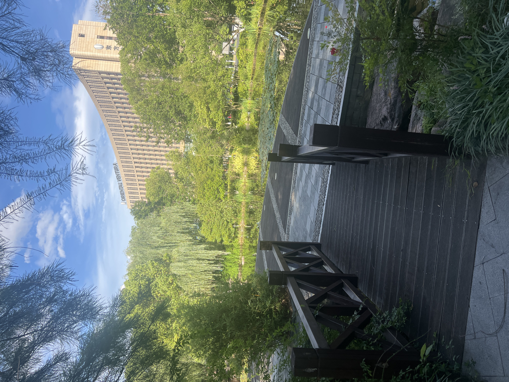 A wooden walkway with dark brown planks and railings, featuring a criss-cross pattern on the railing, leads down towards a body of water. The walkway is situated next to a pond filled with lily pads and surrounded by lush green trees, some of which are weeping willows. In the background, a large, curved, modern building with many windows stands out against a bright blue sky with scattered white clouds. Text on the building reads "ORIENTAL VENTURA" and Chinese characters. The scene is set in a park-like environment, likely in Liandu District, China, given the architecture and vegetation. There are no people visible in the image.