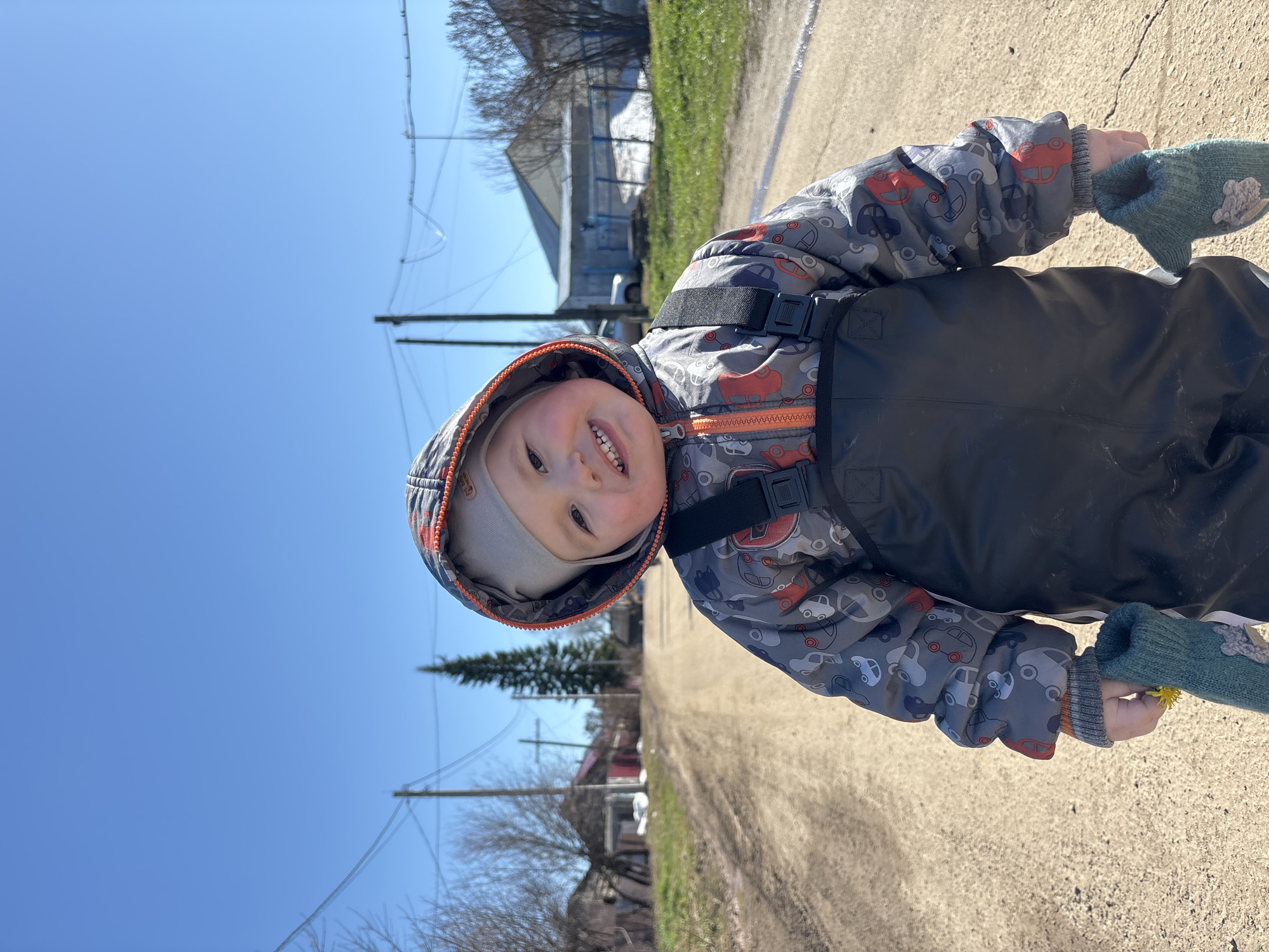 A young child stands outdoors in Фоки, Russia, smiling at the camera. The child wears a hooded jacket with car-themed patterns, black overalls with suspender straps, and green mittens. The child holds a dandelion in their gloved hand. The background shows a dirt road, utility poles with wires, houses, grass, and a clear blue sky.