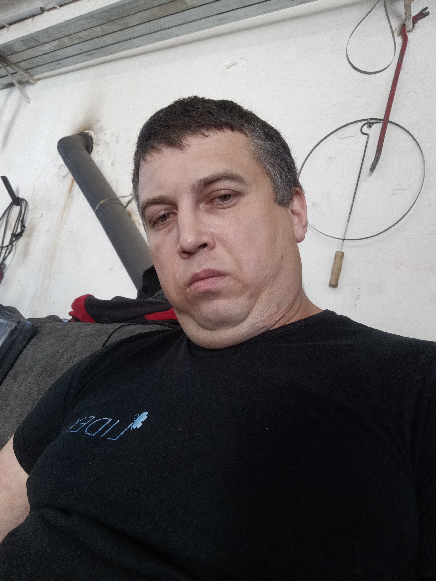 A middle-aged man is depicted in a chest-up, eye-level shot, looking directly into the camera. He has short, dark hair with visible grey strands and a somewhat neutral to slightly somber expression, with his mouth slightly downturned. He has a visible double chin. The man is wearing a black t-shirt that features a light blue design on the left chest, including text that appears to be "FIDEL" or similar, alongside a small butterfly logo.
The setting is an indoor space, likely a workshop, garage, or utility room in Dalovy, Czechia, indicated by the background elements. The wall behind him is light-colored, possibly white or off-white, with a somewhat unfinished texture. In the upper left, exposed metal beams or slats are visible on the ceiling or upper wall, along with hanging electrical wires or cables. A dark, possibly insulated pipe or duct runs diagonally across the upper left side of the background. On the right side of the wall, a red-handled crowbar hangs from a nail, and below it, a circular wire object with a wooden handle is also suspended. A dark, upholstered surface, possibly a couch or seat cushion, is visible behind the man's left shoulder, along with a red and black item, potentially a glove or fabric. The man appears to be seated or reclining.
