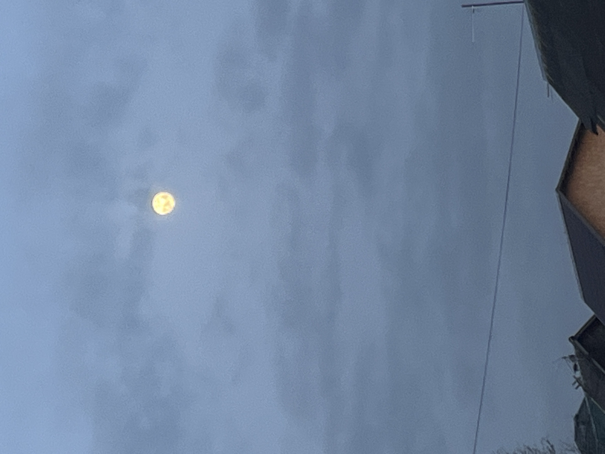 The image shows a daytime view of a cloudy sky in Podolsk, Russia. A full moon is visible through the clouds. Power lines and the corner of a building are in the foreground on the right. No people are visible.