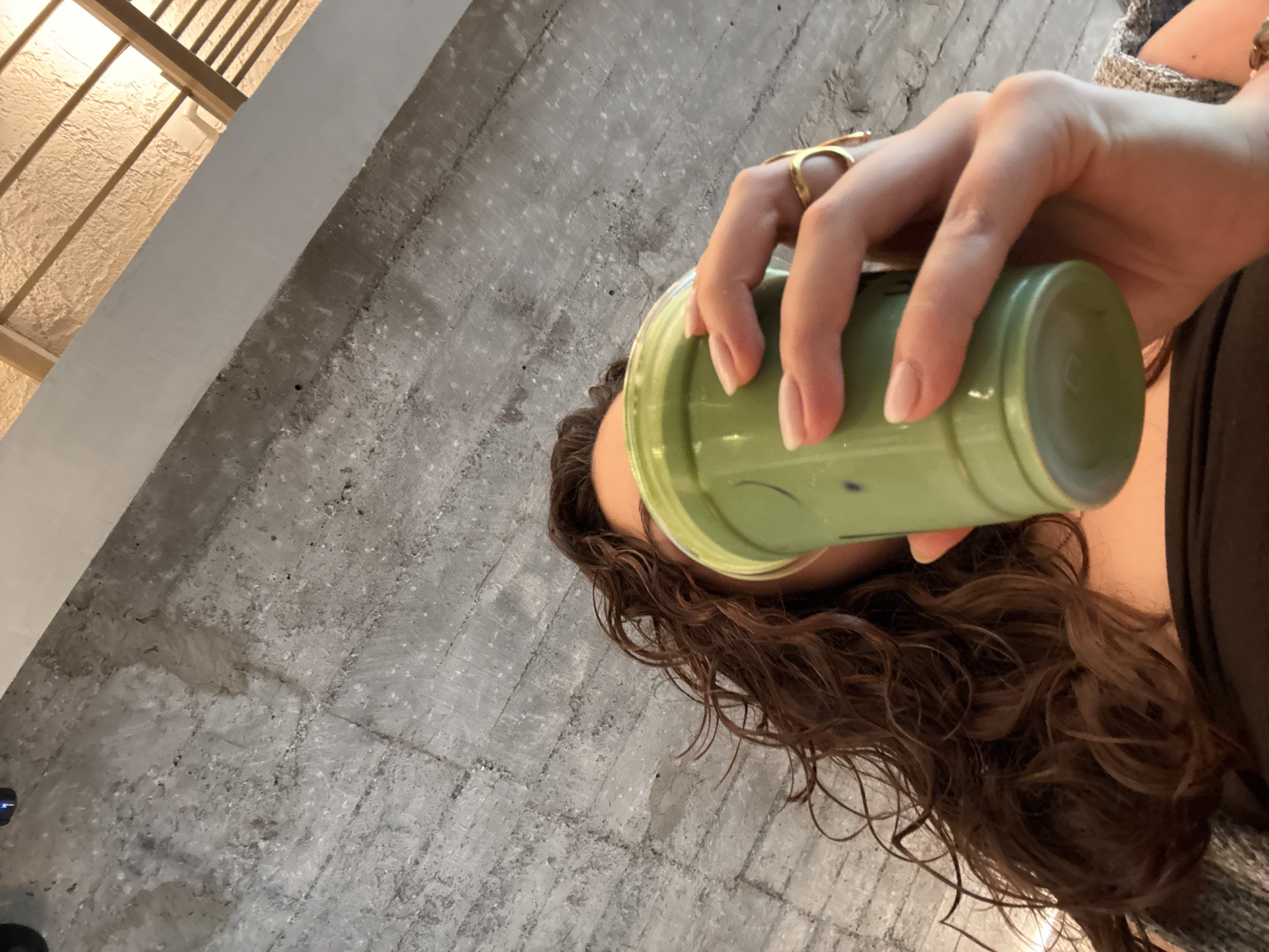 A person with curly brown hair is drinking from a green cup. The person is wearing a gold ring on their right hand. The background is a concrete wall and ceiling. The location is Mexico City, Mexico.