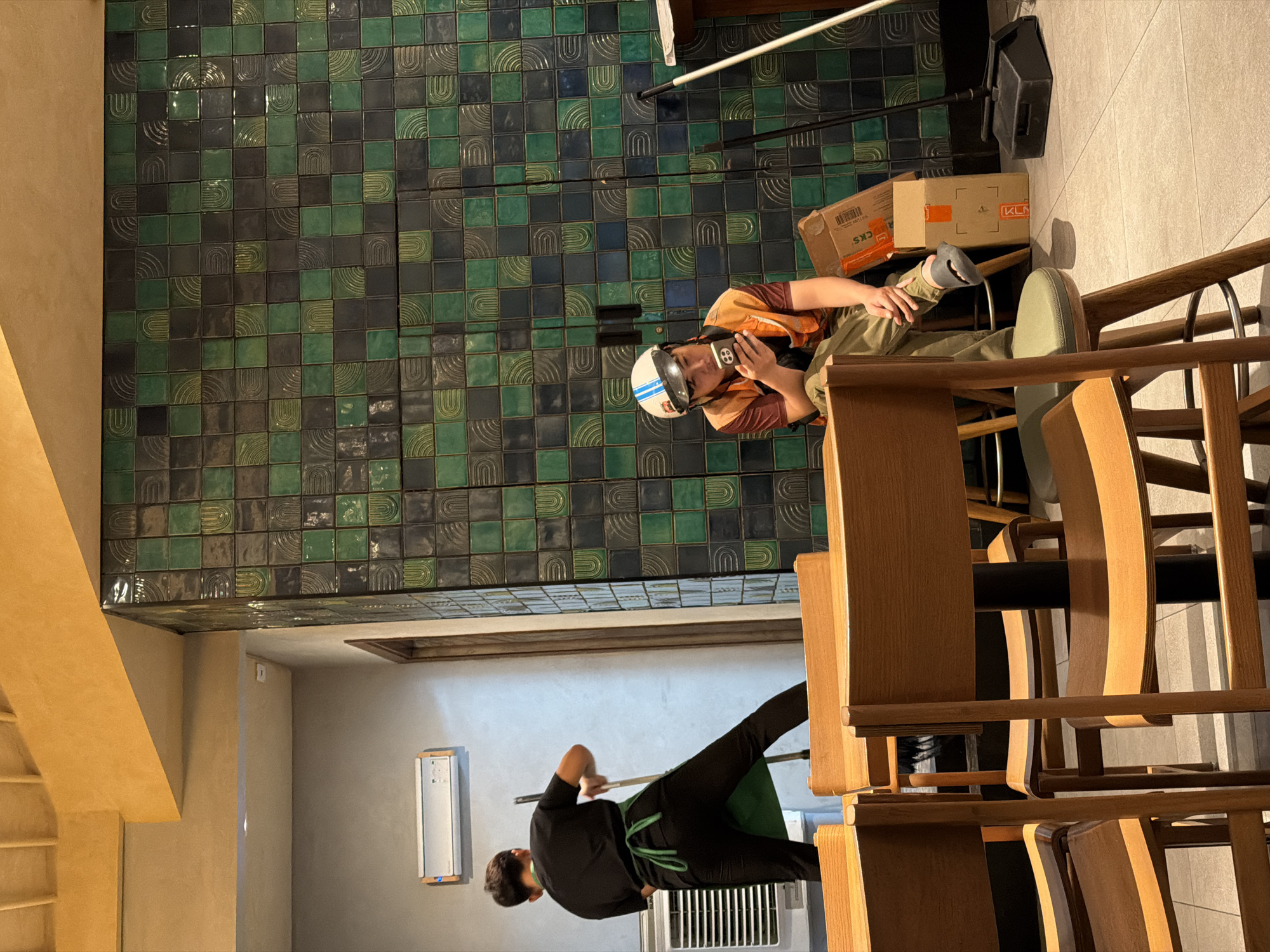 In a cafe in Da Nang, Vietnam, a person wearing a black t-shirt and green apron sweeps the floor with a broom. In the background, a person wearing a safety helmet, orange vest, and black pants sits on a stool, looking at their phone. They are next to a stack of cardboard boxes. The cafe has a wall decorated with green and black tiles. Several wooden chairs are in the foreground.