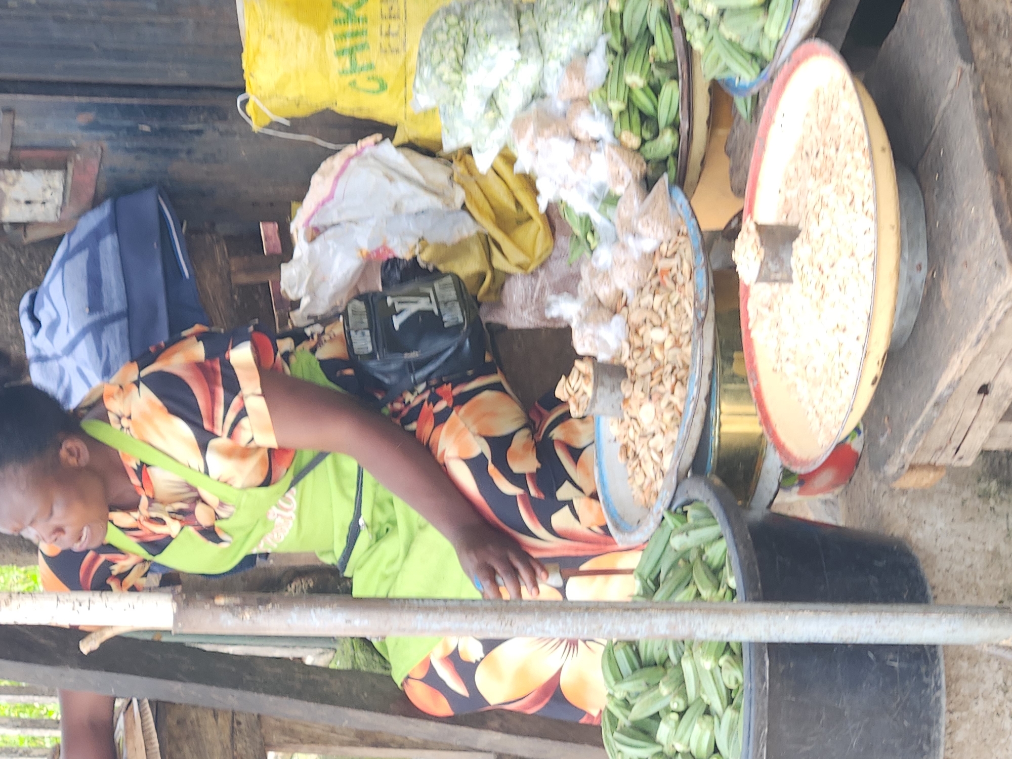 A woman in a green apron and a floral dress sits behind a market stall in Kabong, Nigeria. She is looking up and to the left, with her mouth open as if speaking. In front of her are several bowls of produce and grains. To her left, a large black tub overflows with okra. Next to it, a blue bowl holds more okra. In the center, a metal bowl contains what appears to be dried seeds or nuts. To the right, a yellow bag is partially visible, with the word "CHIK" printed on it. Several plastic bags of produce are also stacked in the background. The scene is lit by natural light, suggesting it is daytime.