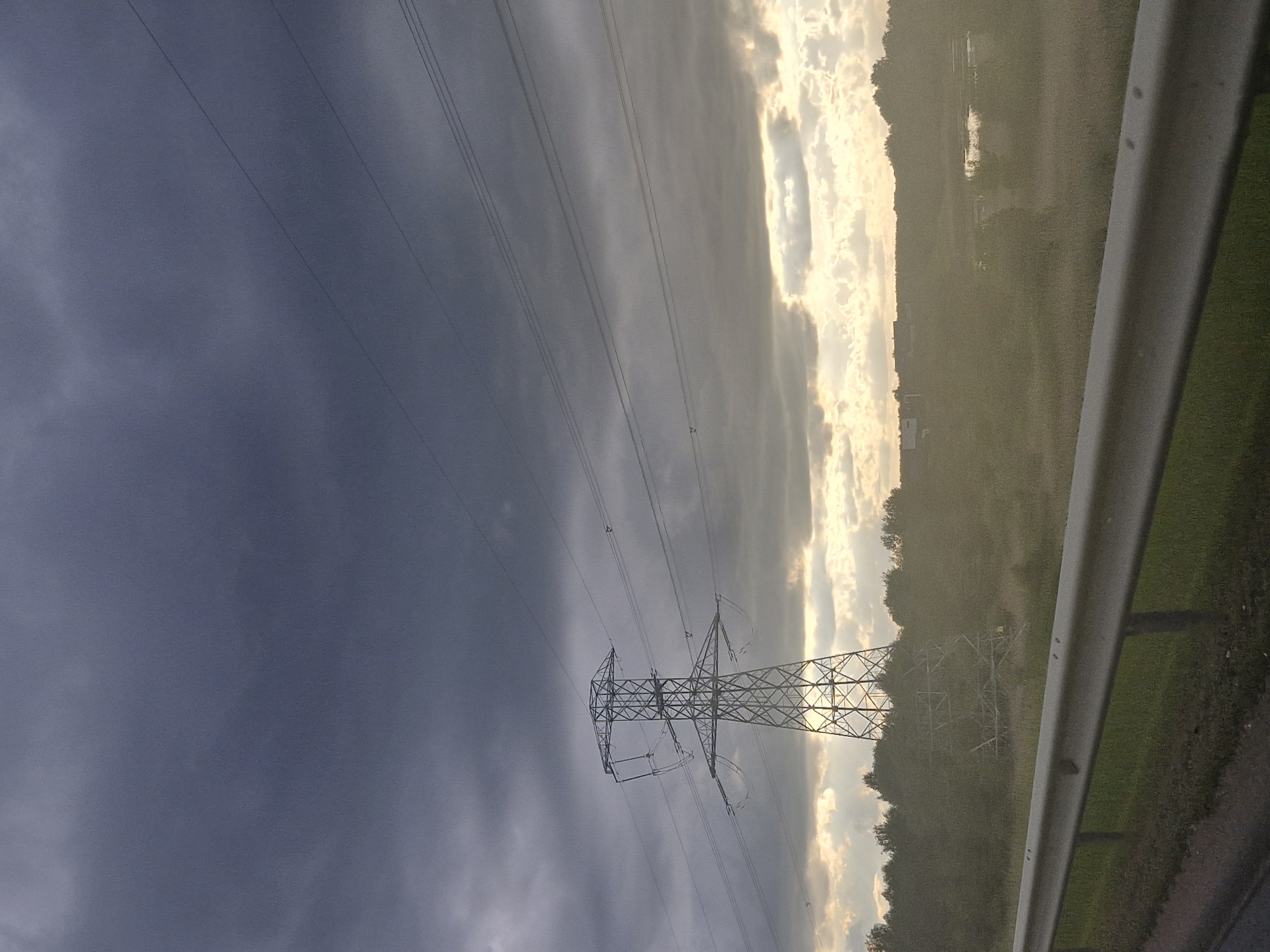 The image displays an outdoor, semi-rural landscape in Unknown, Unknown, under an overcast sky with breaks of sunlight, suggesting either dawn or dusk. A large, lattice-steel electrical transmission tower (pylon) is prominently positioned left of center, its structure silhouetted against a bright, diffused light source behind a break in the clouds. Multiple power lines extend horizontally from the pylon's cross-arms across the upper and middle portions of the frame. In the immediate foreground, a silver-colored guardrail extends horizontally across the lower frame, backed by green grass and a dark, indistinct road surface. The midground consists of a wide, hazy field of green-brown vegetation, bordered by a treeline. A small body of water and several distant, low-rise structures are visible further back, partially obscured by atmospheric haze. The upper sky is dominated by dark, heavy grey clouds, transitioning to lighter, illuminated clouds towards the horizon. No people are present.