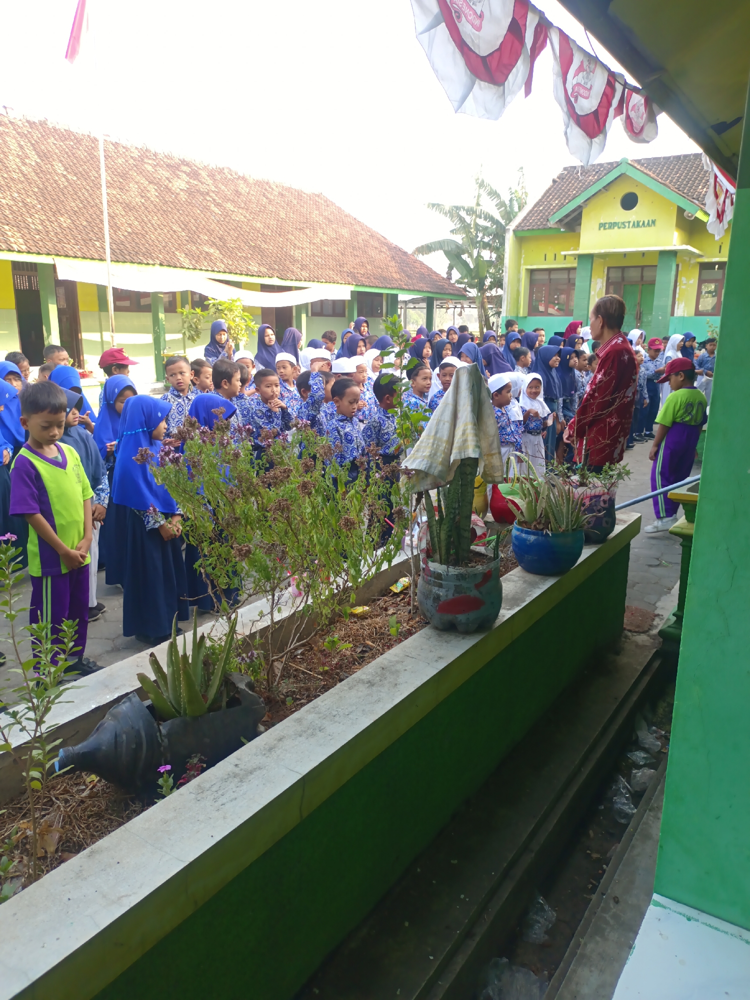 A group of children in school uniforms are gathered outdoors in front of a yellow building with "PERPUSTAKAAN" (Library) written on it. Many of the children are wearing blue shirts and pants or hijabs. A few adults are also present, including one in a red and white patterned shirt. The children appear to be looking towards a woman in a red top who is facing them. The scene suggests an outdoor school activity or assembly. The environment includes trees, a tiled roof building, and decorative plants in planters made from repurposed materials. The image appears to have been taken in Sarimulyo, Indonesia, as indicated by the presence of Indonesian text on the building and the style of the school uniforms.